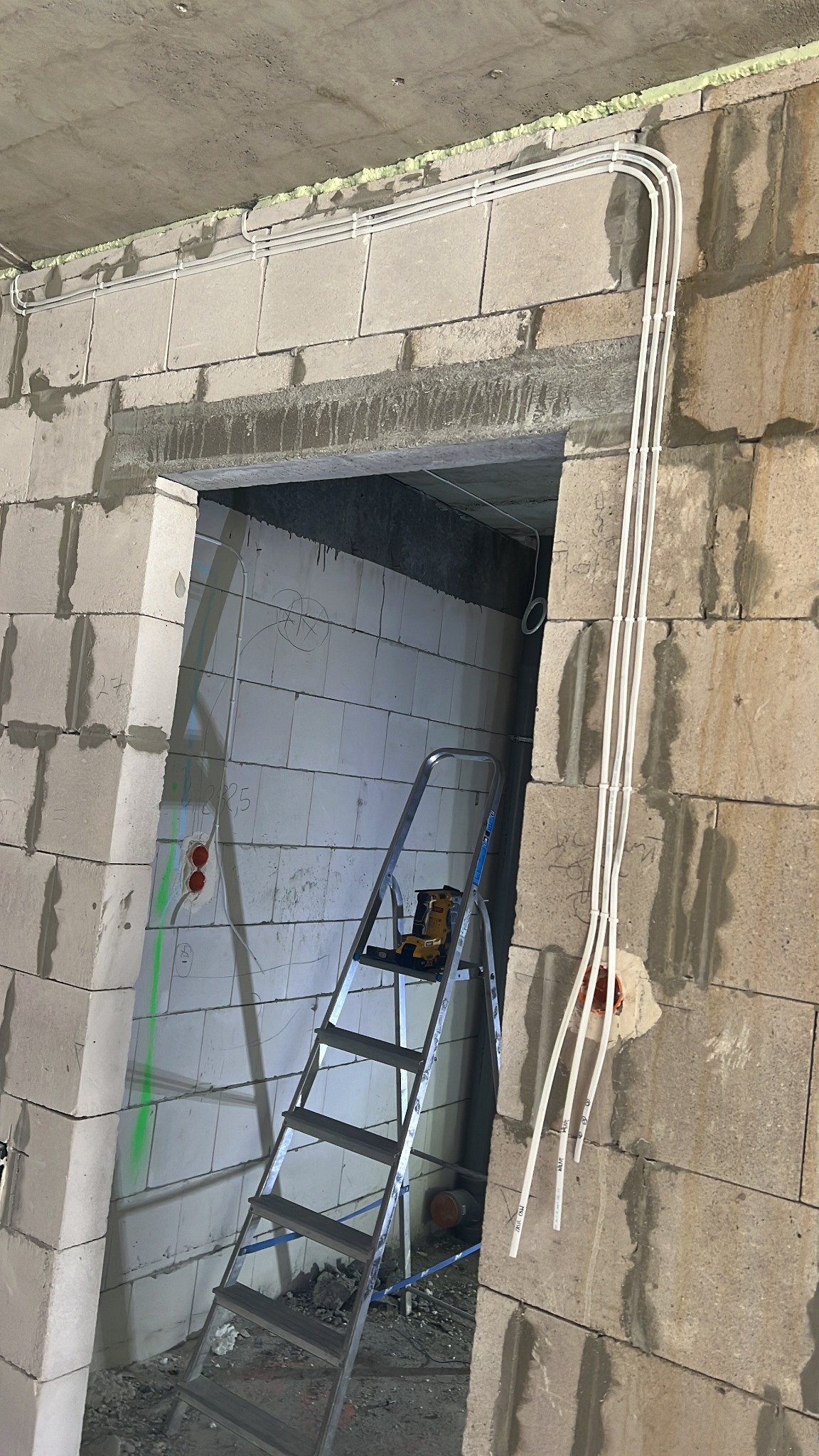 Białe przewody elektryczne ułożone wzdłuż surowej ściany z pustaków, widoczne puszki instalacyjne i drabina. Etap prac instalacyjnych wewnątrz budynku.