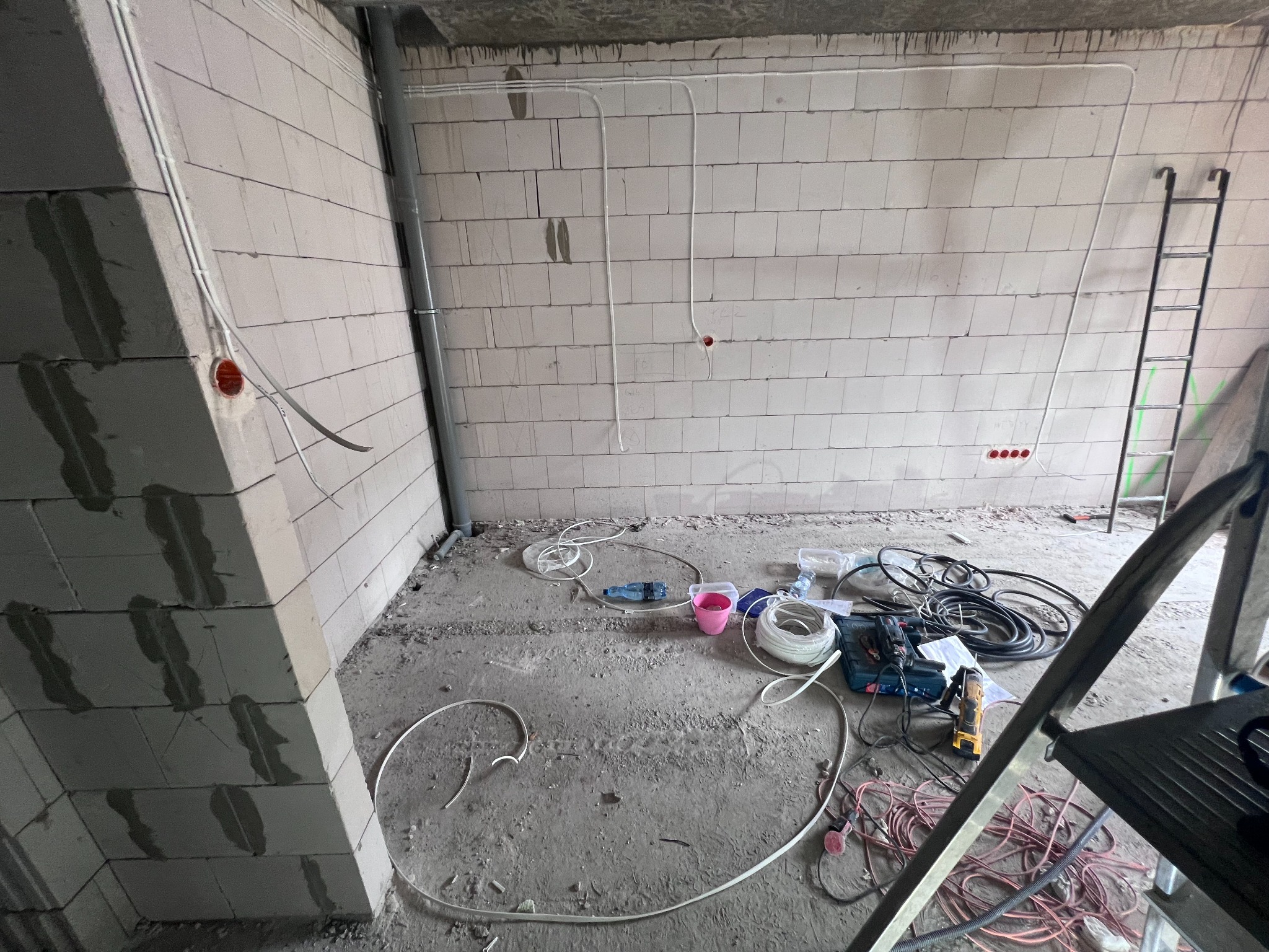 Instalacja elektryczna w surowym wnętrzu: białe kable prowadzone po ścianach z betonu komórkowego, narzędzia i drabina w tle. Etap prac przed wykończeniem.