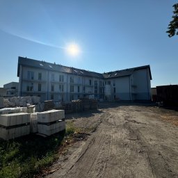 CHUKHACH GROUP POLSKA (CGP) SPÓŁKA Z OGRANICZONĄ ODPOWIEDZIALNOŚCIĄ - Nowoczesny budynek wielorodzinny w trakcie budowy, widok z dołu na jasną elewację i materiały budowlane na placu. Słoneczny dzień.