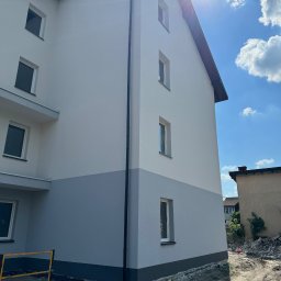 CHUKHACH GROUP POLSKA (CGP) SPÓŁKA Z OGRANICZONĄ ODPOWIEDZIALNOŚCIĄ - Dwukolorowa elewacja budynku mieszkalnego w słoneczny dzień. Górna część biała, dolna szara. Widoczne okna i rynna. Na pierwszym planie fragment rusztowania.