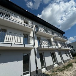 CHUKHACH GROUP POLSKA (CGP) SPÓŁKA Z OGRANICZONĄ ODPOWIEDZIALNOŚCIĄ - Nowoczesny budynek wielorodzinny z szarą elewacją i czarnymi balkonami, uchwycony pod kątem z widocznym niebem i chmurami. Minimalistyczny design.