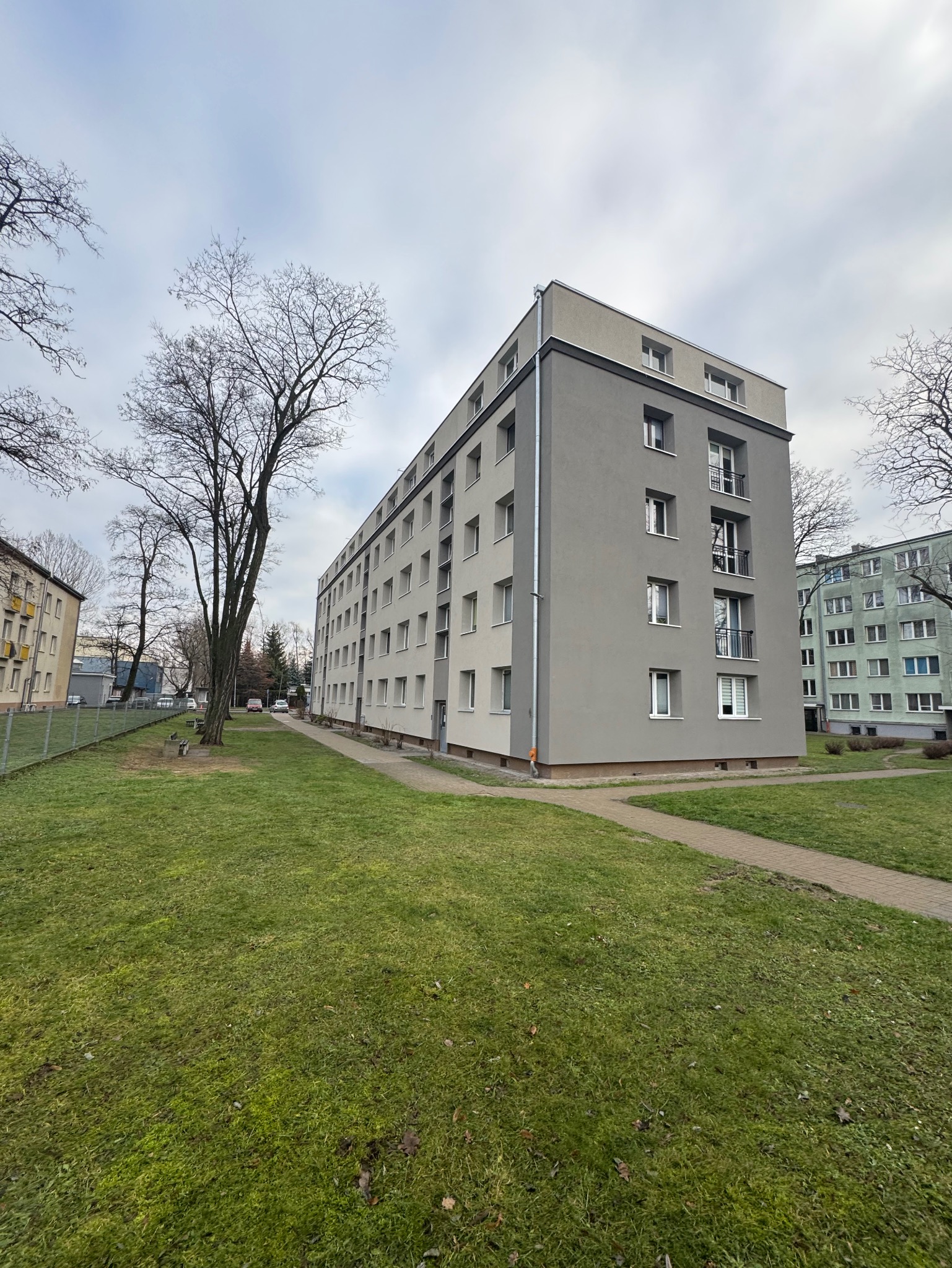 Szary budynek wielorodzinny po renowacji, widok z trawnika. Nowoczesna elewacja z balkonami i oknami. Minimalistyczny design, spokojna kolorystyka, jesienny krajobraz.