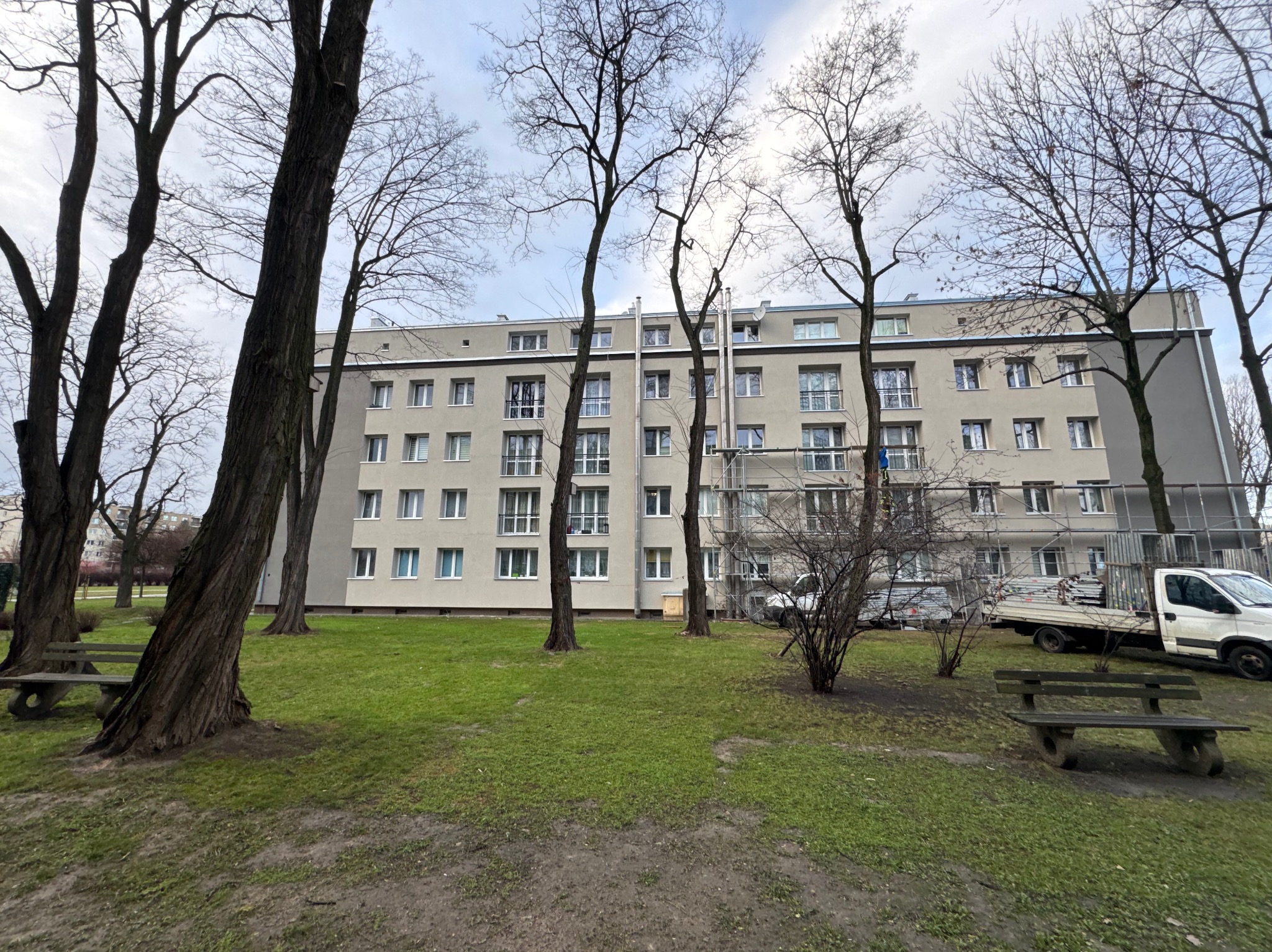 Wielorodzinny budynek z beżową elewacją, częściowo osłonięty rusztowaniami. Widoczne drzewa i trawnik. Na drugim planie zaparkowany samochód dostawczy.