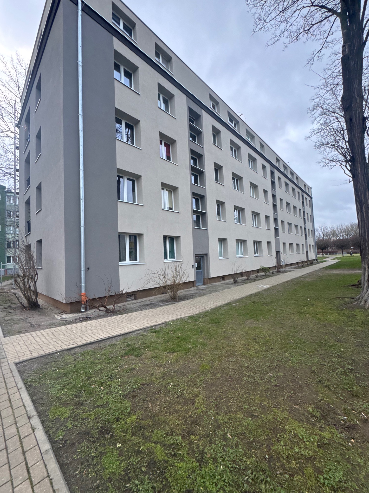 Odnowiony blok mieszkalny z szarą elewacją i nową rynną, widoczny z poziomu chodnika i trawnika. Minimalistyczny styl, pochmurne niebo w tle.
