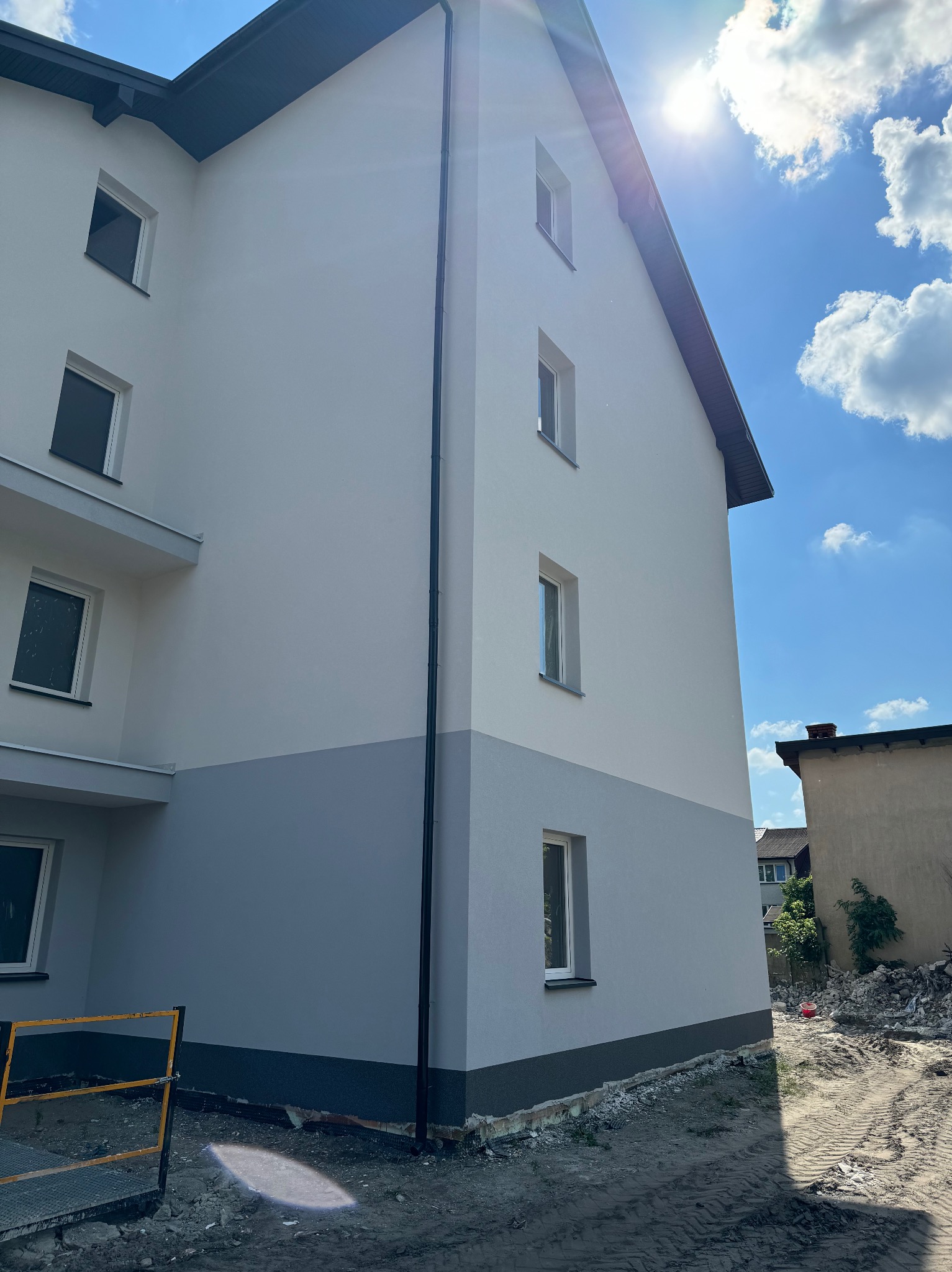 Dwukolorowa elewacja budynku mieszkalnego w słoneczny dzień. Górna część biała, dolna szara. Widoczne okna i rynna. Na pierwszym planie fragment rusztowania.