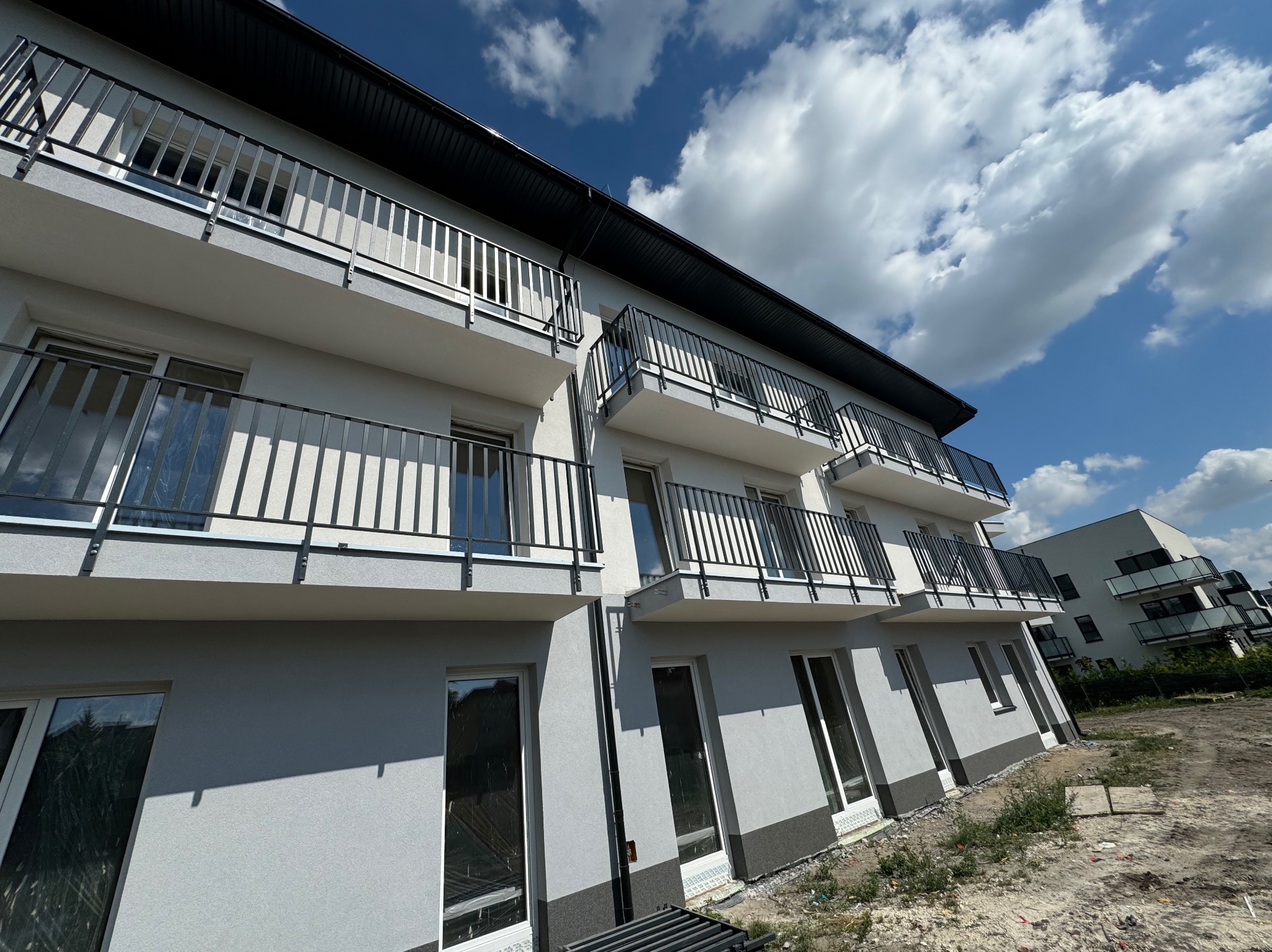 Nowoczesny budynek wielorodzinny z szarą elewacją i czarnymi balkonami, uchwycony pod kątem z widocznym niebem i chmurami. Minimalistyczny design.