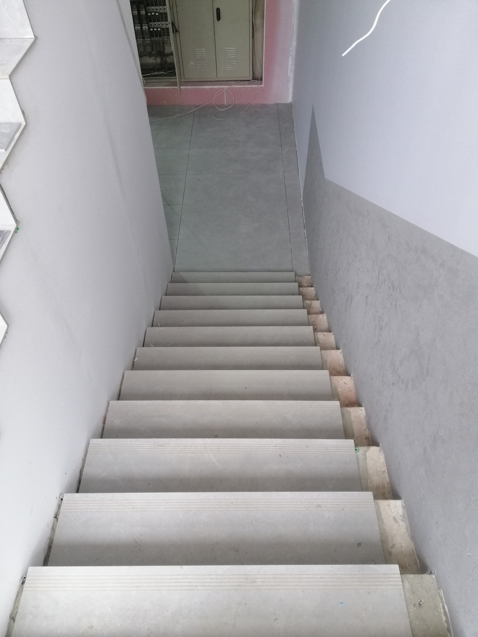 Wykończone schody w szarościach, widok z góry. Szare płytki na stopniach, szare ściany, widoczna instalacja elektryczna w tle.