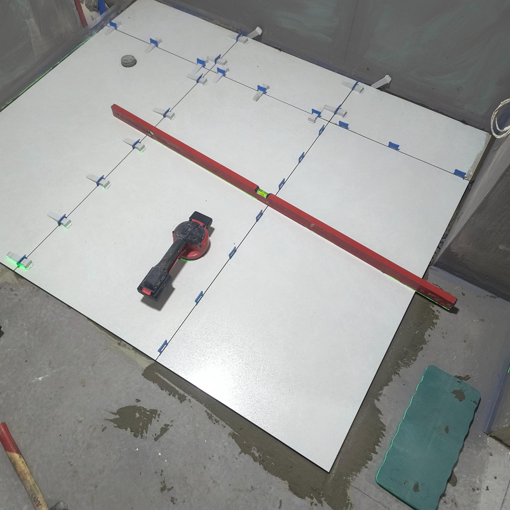 Układanie białych płytek podłogowych z użyciem poziomicy laserowej i klipsów poziomujących. Widoczne narzędzia glazurnicze i fragment podłogi z zaprawą.