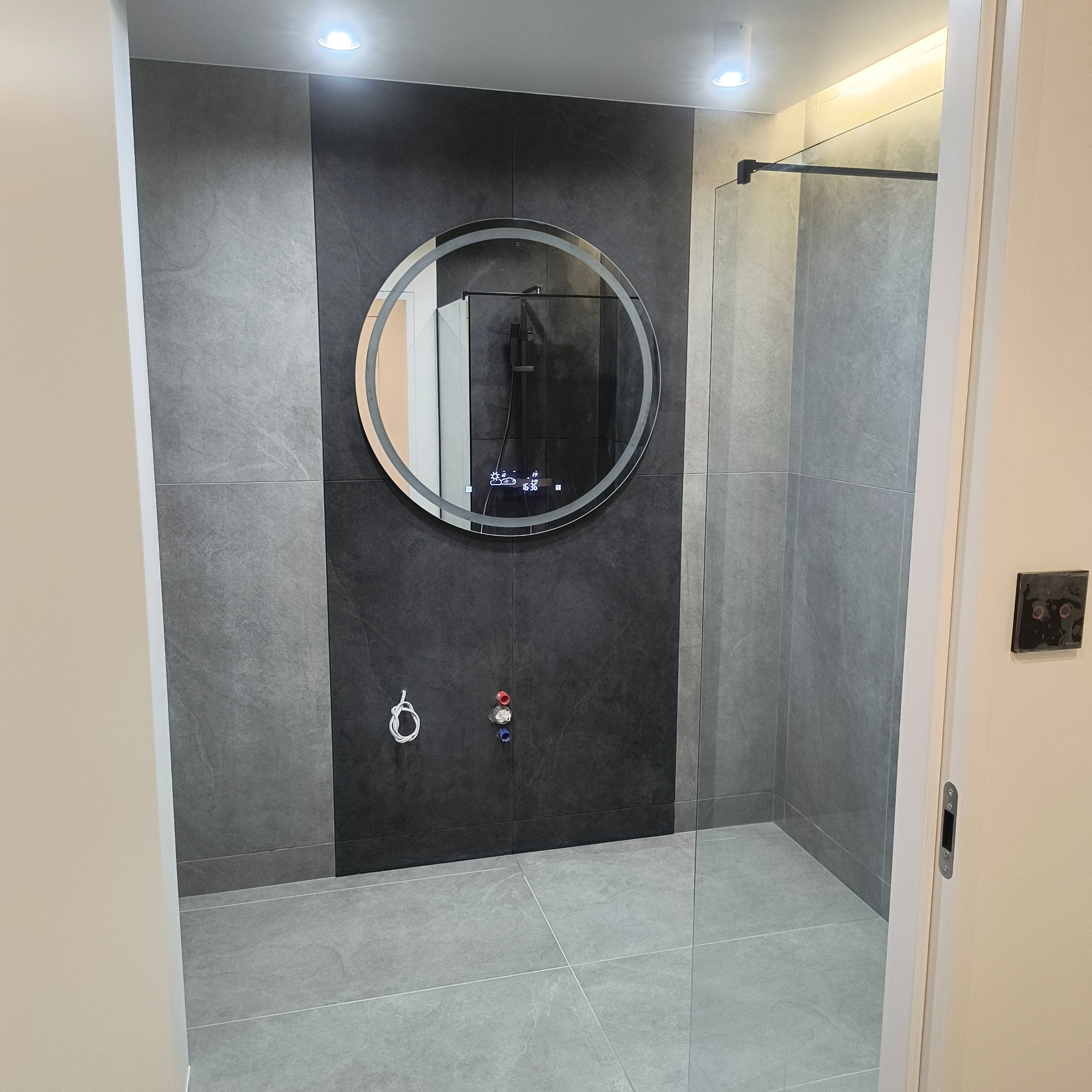 Nowoczesna łazienka z szarą glazurą, okrągłym lustrem z podświetleniem LED i szklaną kabiną prysznicową. Widoczne przyłącza wodne i minimalistyczny design.