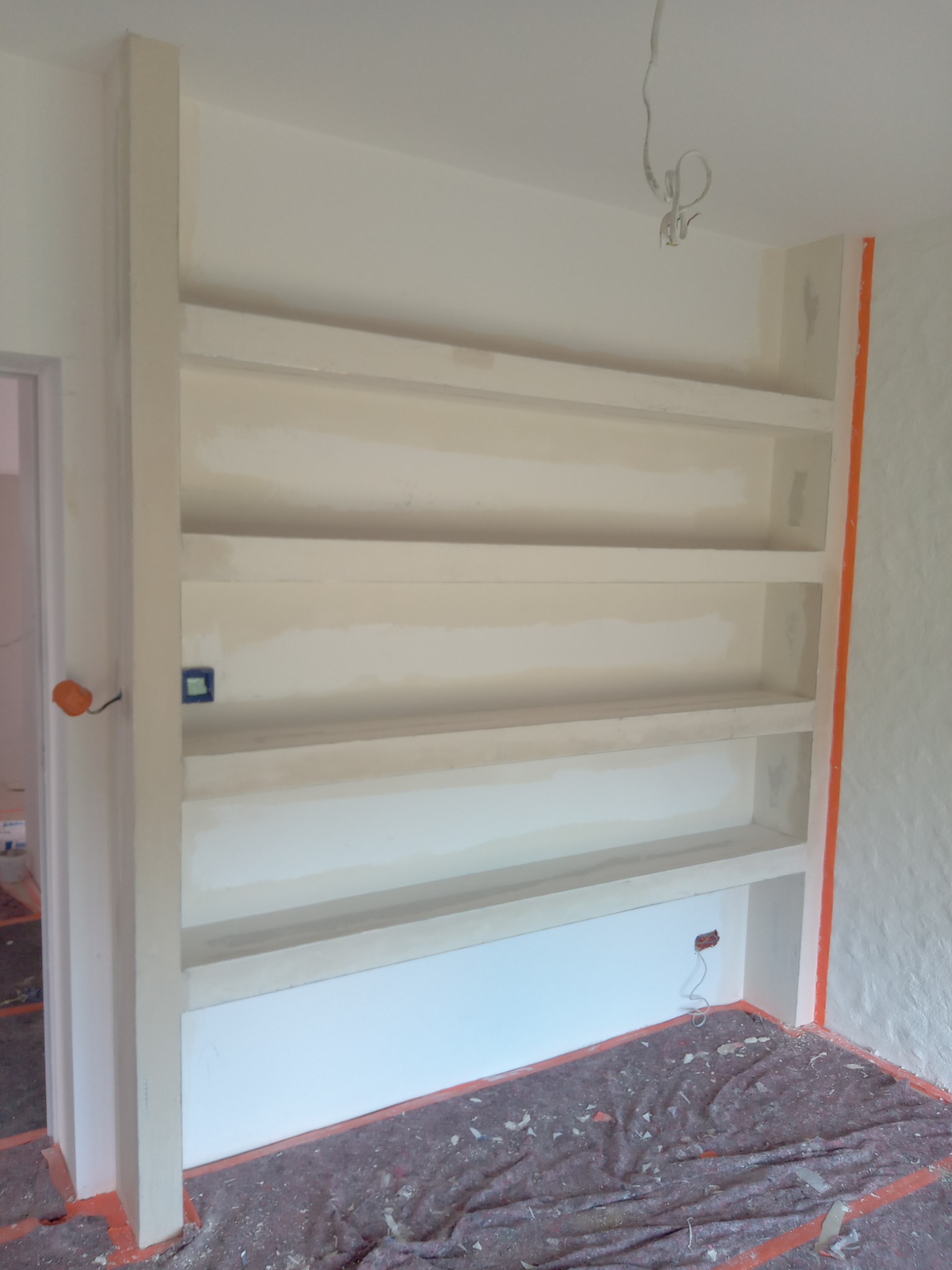 Wnęka z półkami w trakcie szpachlowania, zabezpieczona podłoga, widoczne puszki elektryczne. Element wykończenia wnętrza w trakcie prac remontowych.