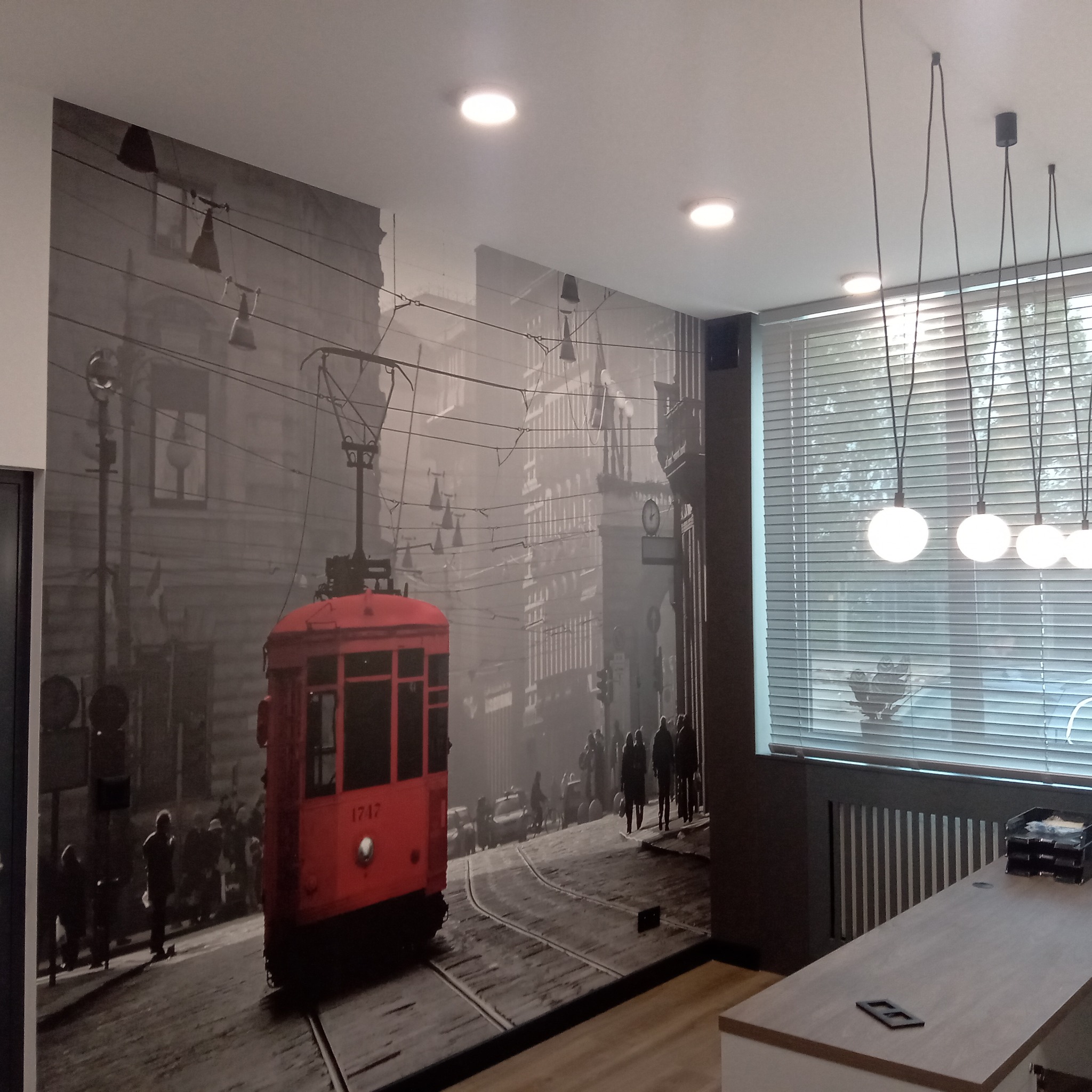 Fototapeta w biurze z motywem retro: czerwony tramwaj na tle szarego miasta. Nowoczesne lampy kuliste i żaluzje w tle.