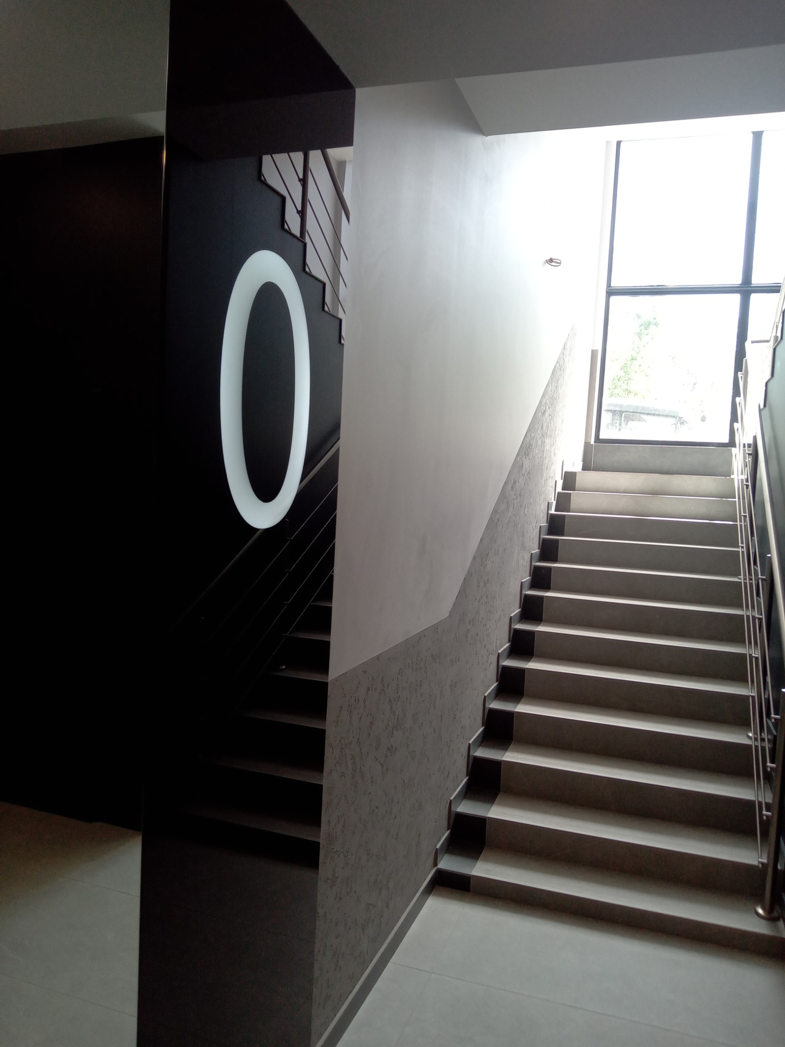 Klatka schodowa w nowoczesnym budynku. Ciemna ściana z podświetlonym numerem '0', jasne schody z metalową barierką prowadzą do okna.