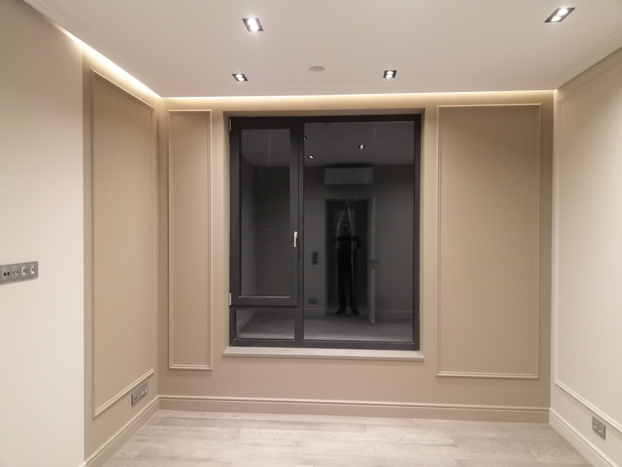 Eleganckie wykończenie wnętrza z panelami ściennymi w odcieniach beżu, oświetleniem LED podsufitowym i nowoczesnym oknem. Widoczny parkiet i gniazdka elektryczne.