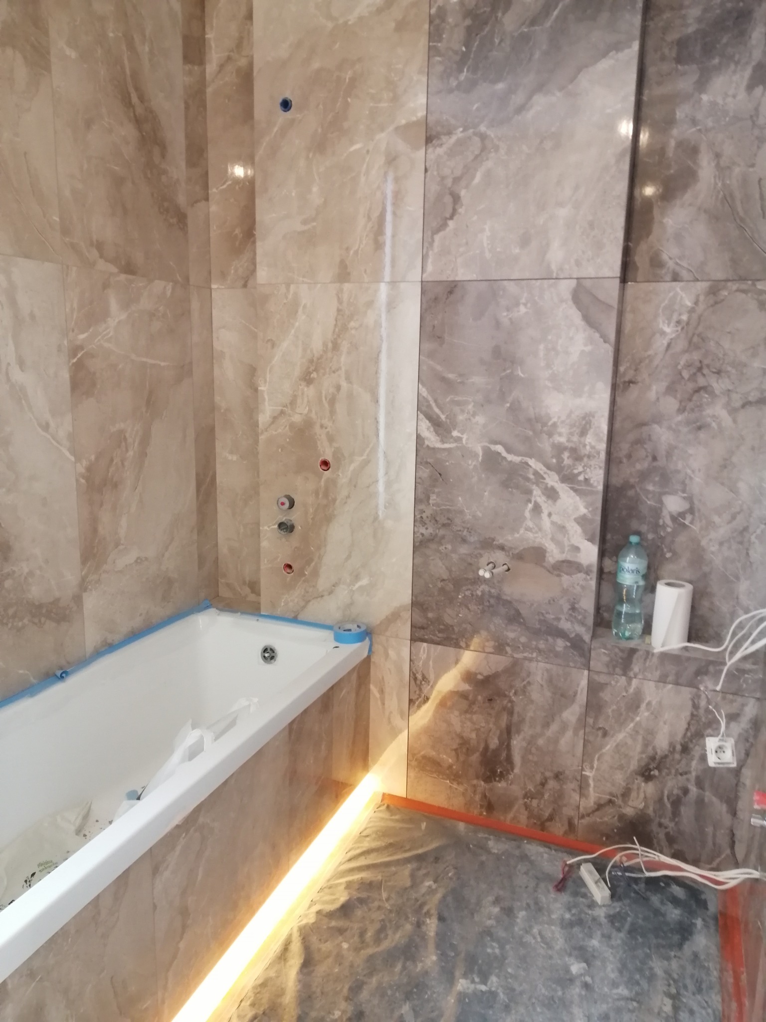 Łazienka w trakcie wykańczania: wanna obłożona płytkami imitującymi marmur, widoczne instalacje wodne i elektryczne, podświetlenie LED, butelka wody i rolka papieru.