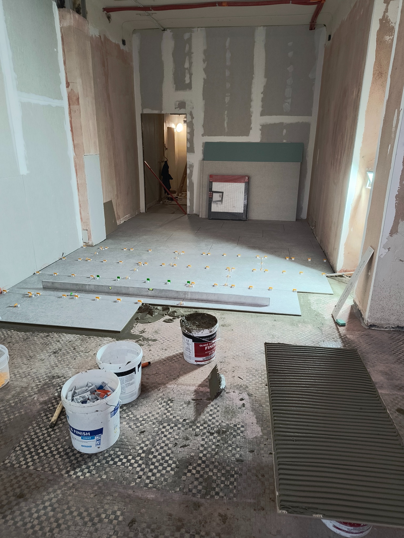 Układanie szarych płytek podłogowych z użyciem poziomicy i krzyżyków dystansowych. Widoczne wiadra z klejem i płytka z nałożonym klejem. Etap prac wykończeniowych.