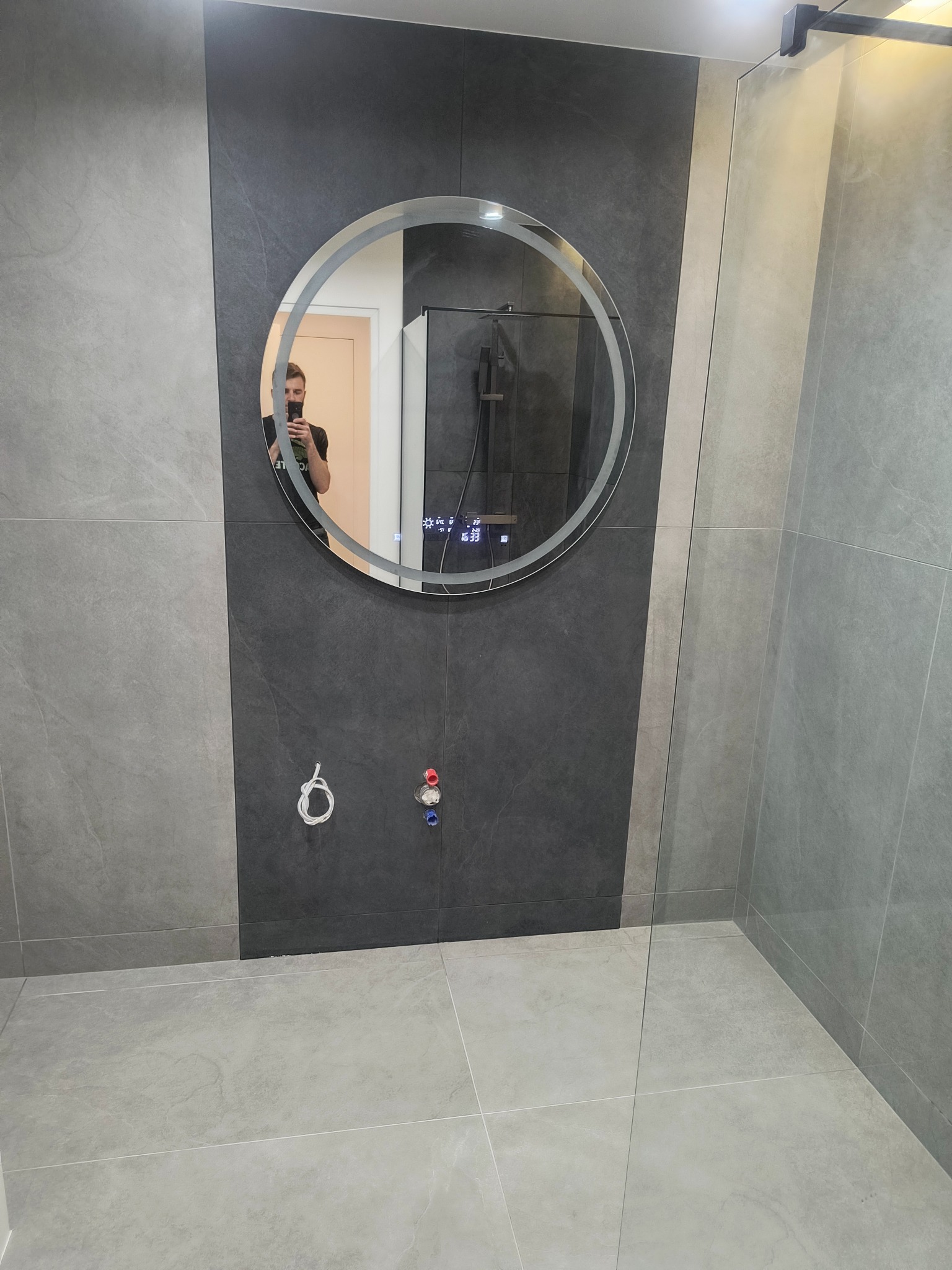 Nowoczesna łazienka w Łodzi z szarymi płytkami, okrągłym lustrem z podświetleniem i szklaną kabiną prysznicową. Widoczne przyłącza hydrauliczne.