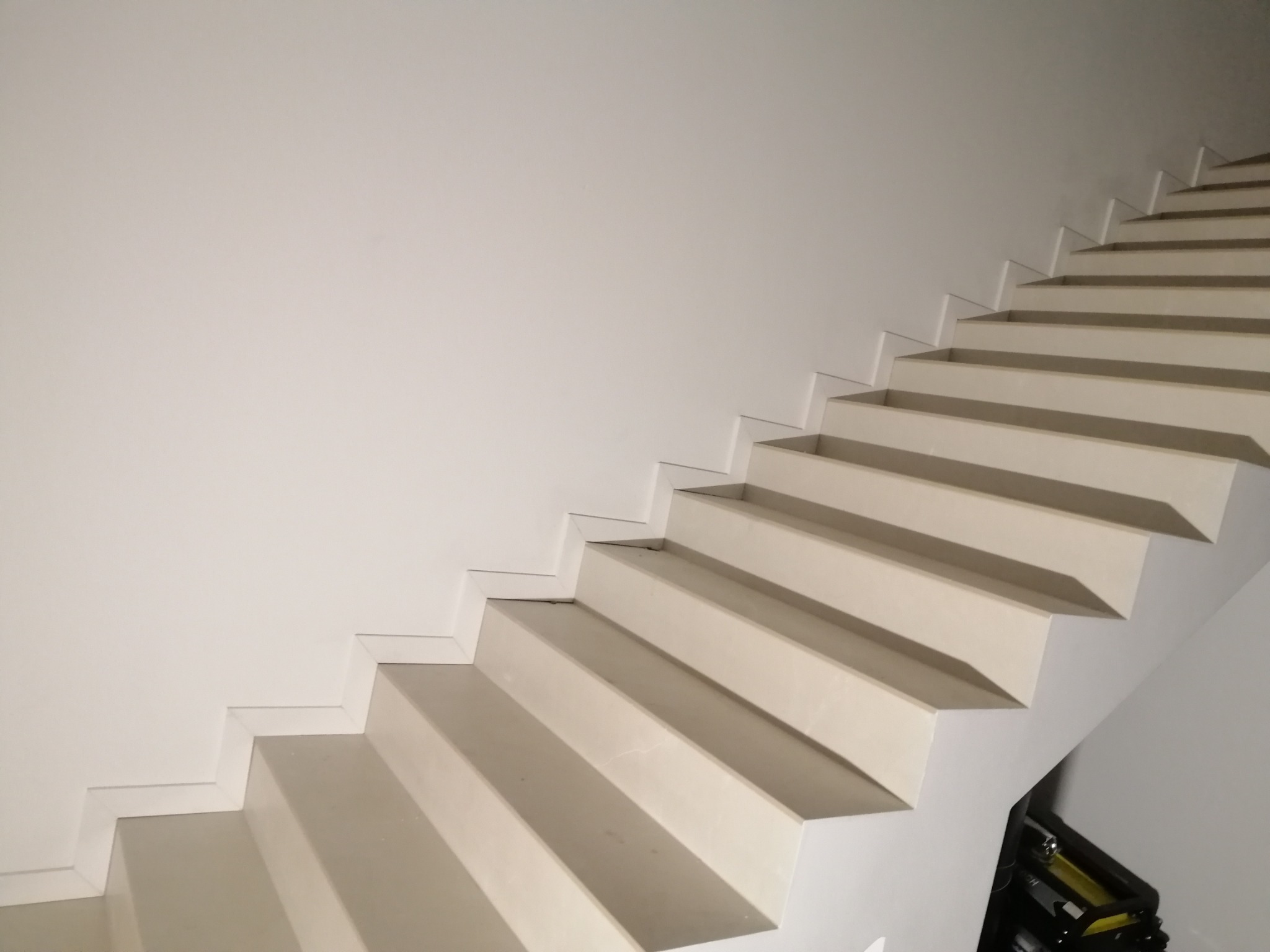 Minimalistyczne, białe schody wspornikowe w nowoczesnym wnętrzu. Widoczne geometryczne detale i subtelne oświetlenie. W tle fragment ściany i niezidentyfikowany przedmiot.