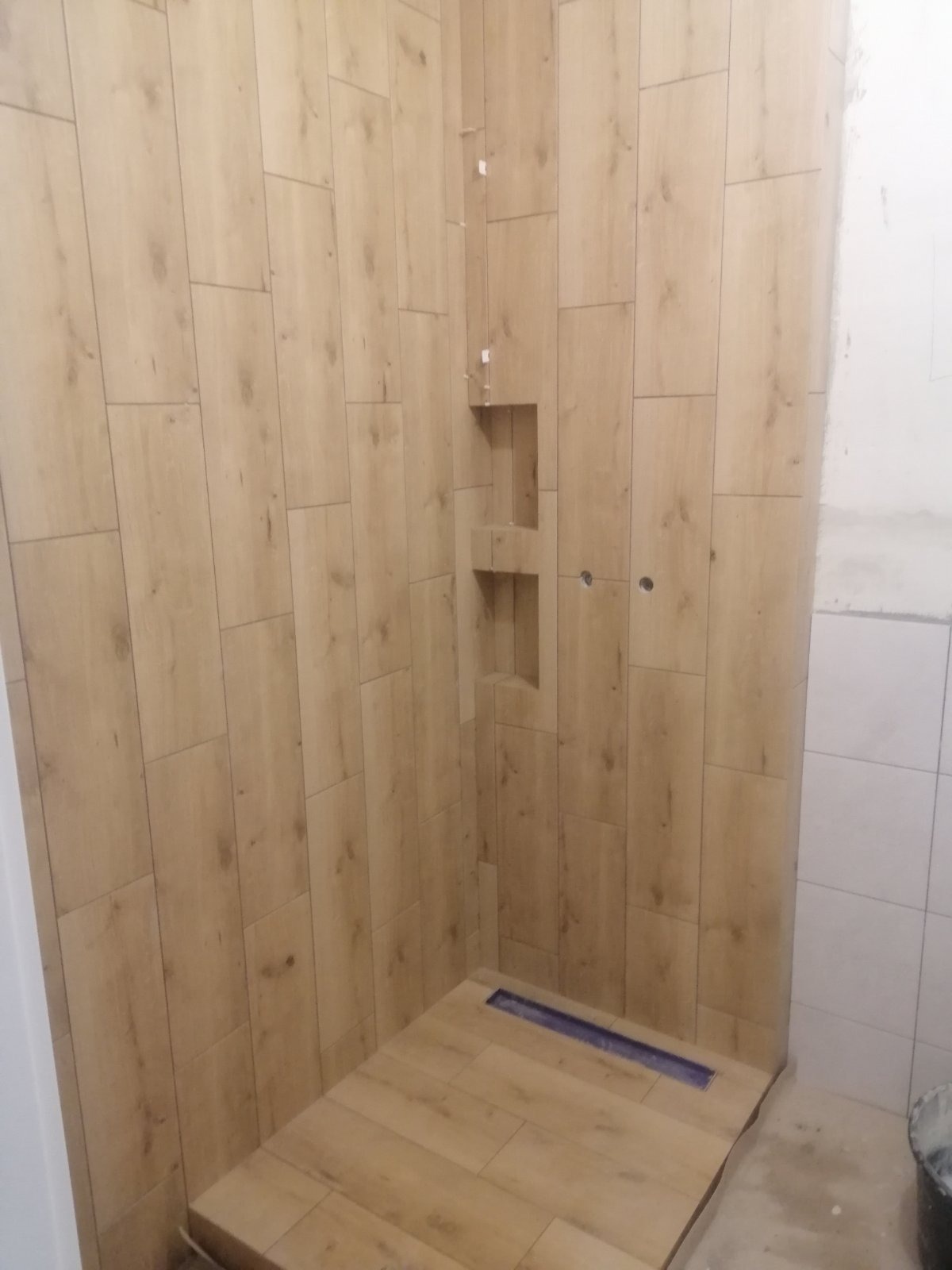 Wykończona kabina prysznicowa z drewnopodobnymi płytkami, wnęką na kosmetyki i odpływem liniowym. Częściowo widoczne białe płytki na ścianie.