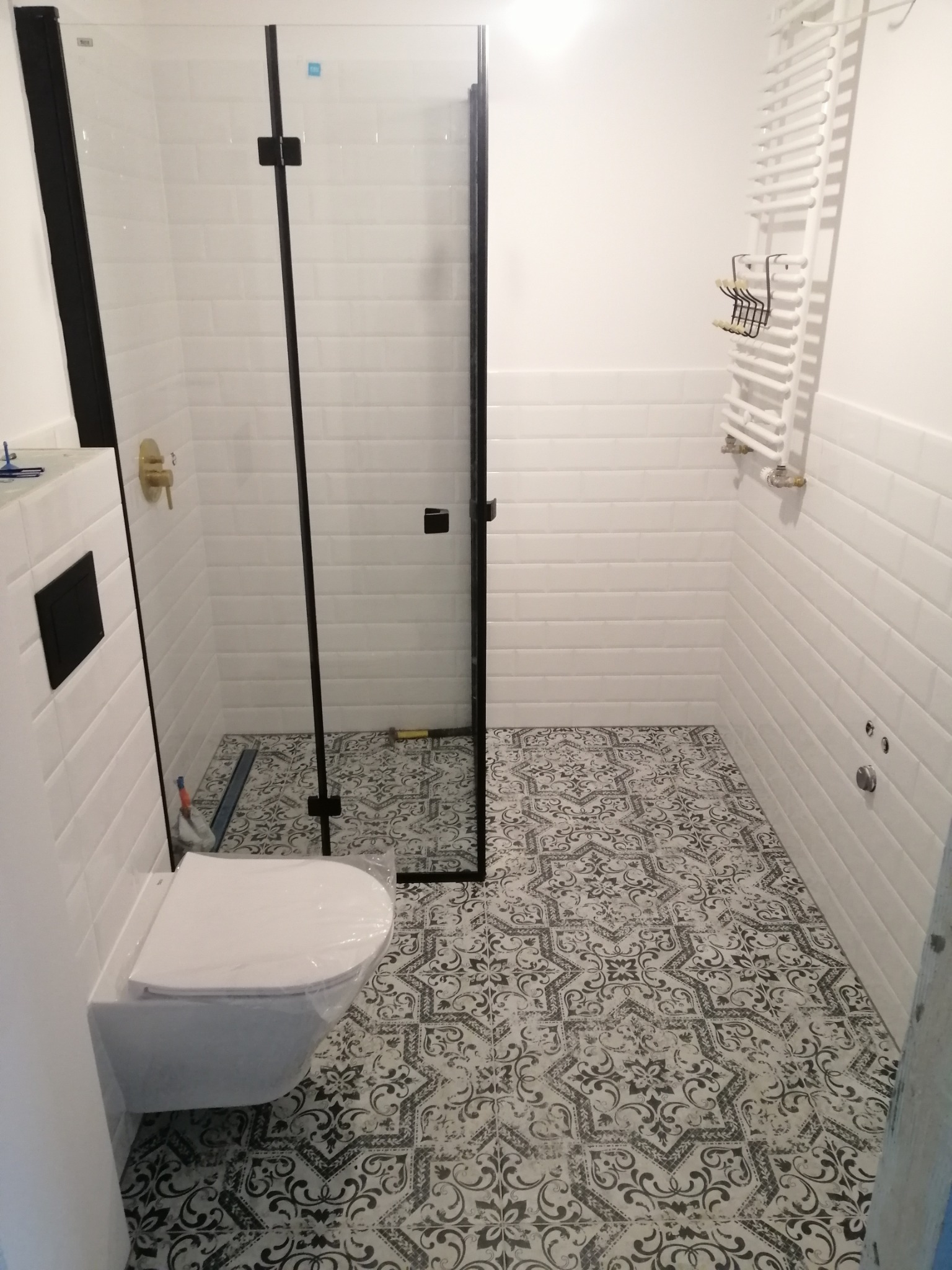 Wykończona łazienka z kabiną prysznicową typu walk-in, białe płytki na ścianach, wzorzysta podłoga. Umywalka i grzejnik drabinkowy. Widoczne narzędzia.