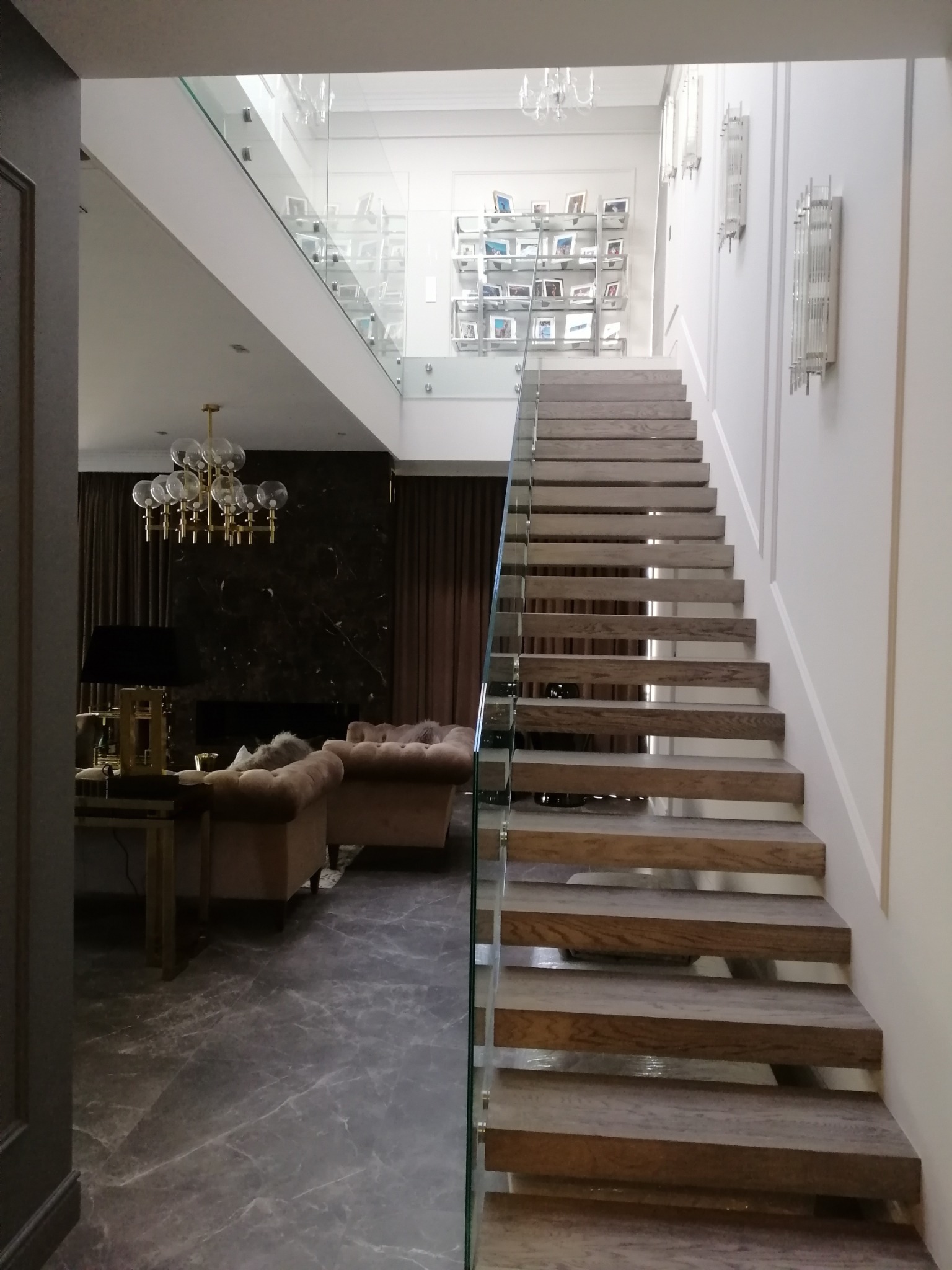 Nowoczesne schody drewniane z szklaną balustradą w przestronnym wnętrzu domu z marmurową podłogą i eleganckim wystrojem salonu w tle.