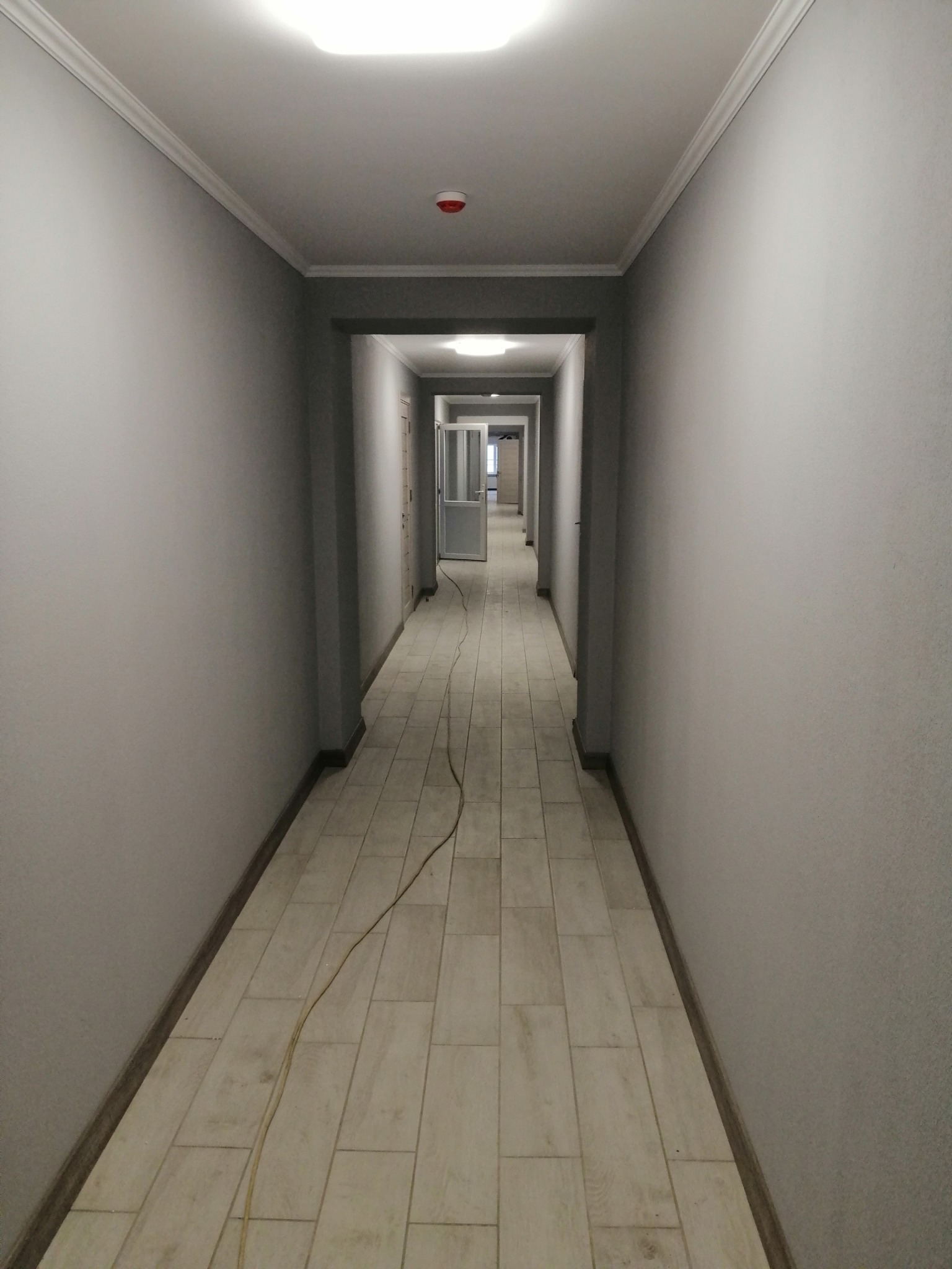 Długi, wąski korytarz po remoncie z szarymi ścianami, jasną podłogą z płytek imitujących drewno i białym wykończeniem sufitu. Widok w głąb korytarza.