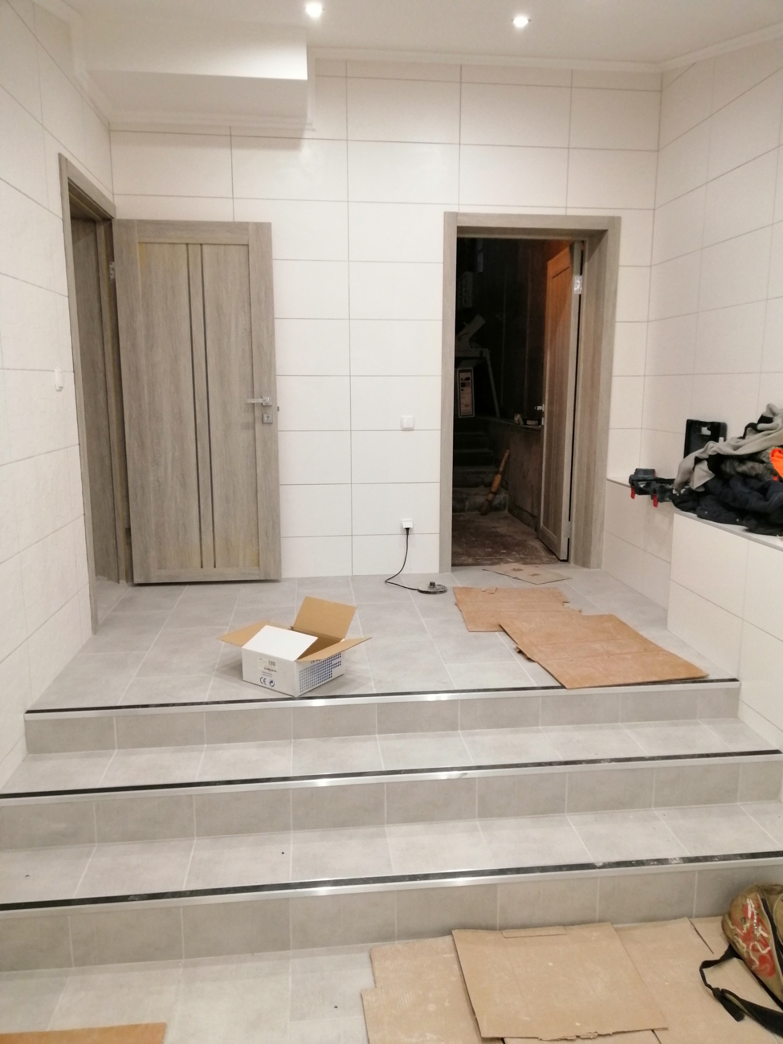 Wykończone schody z płytek w jasnej tonacji, z widocznymi listwami. W tle drzwi i białe płytki na ścianach. Na podłodze kartony i pudełko.