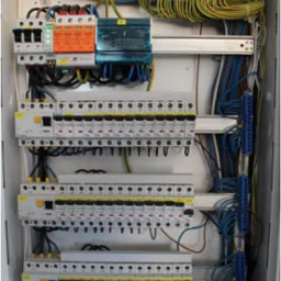 Rozdzielnia elektryczna z widocznymi zabezpieczeniami, przewodami o różnych kolorach i listwami zaciskowymi. Widok z bliska na wnętrze skrzynki elektrycznej.