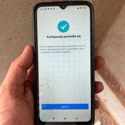 VoltMaster PAWEŁ MIŚKIEWICZ - Ekran smartfona w dłoni z komunikatem o pomyślnej konfiguracji. Widoczny stan sieci 5G. Tło to surowa ściana w trakcie prac wykończeniowych.
