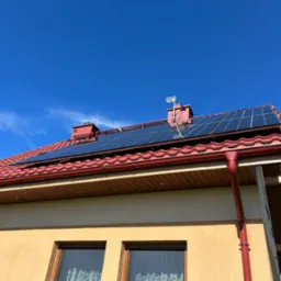 Panele słoneczne na dachu domu z czerwoną dachówką, widoczne okna i błękitne niebo. Instalacja w słoneczny dzień, antena na kominie.