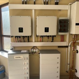 VoltMaster PAWEŁ MIŚKIEWICZ - Kompletna instalacja fotowoltaiczna w pomieszczeniu: dwa inwertery FOX, dwa akumulatory Miellec, rozdzielnice i okablowanie na beżowej ścianie, obok okna i bojlera.