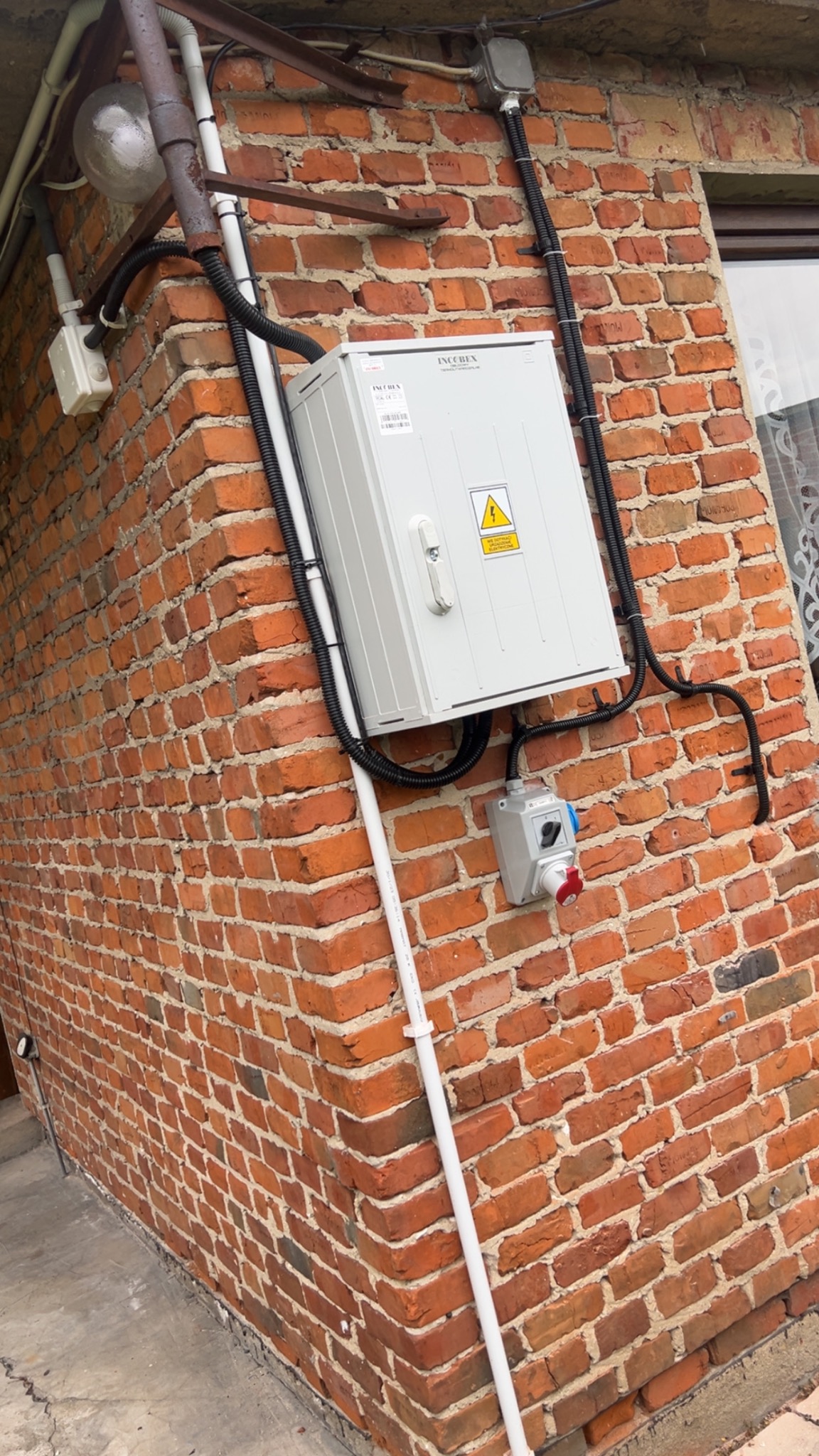 Szara skrzynka elektryczna z symbolem ostrzegawczym, zamontowana na czerwonej, ceglanej ścianie budynku. Widoczne przewody w czarnych peszlach i biała rura PCV.