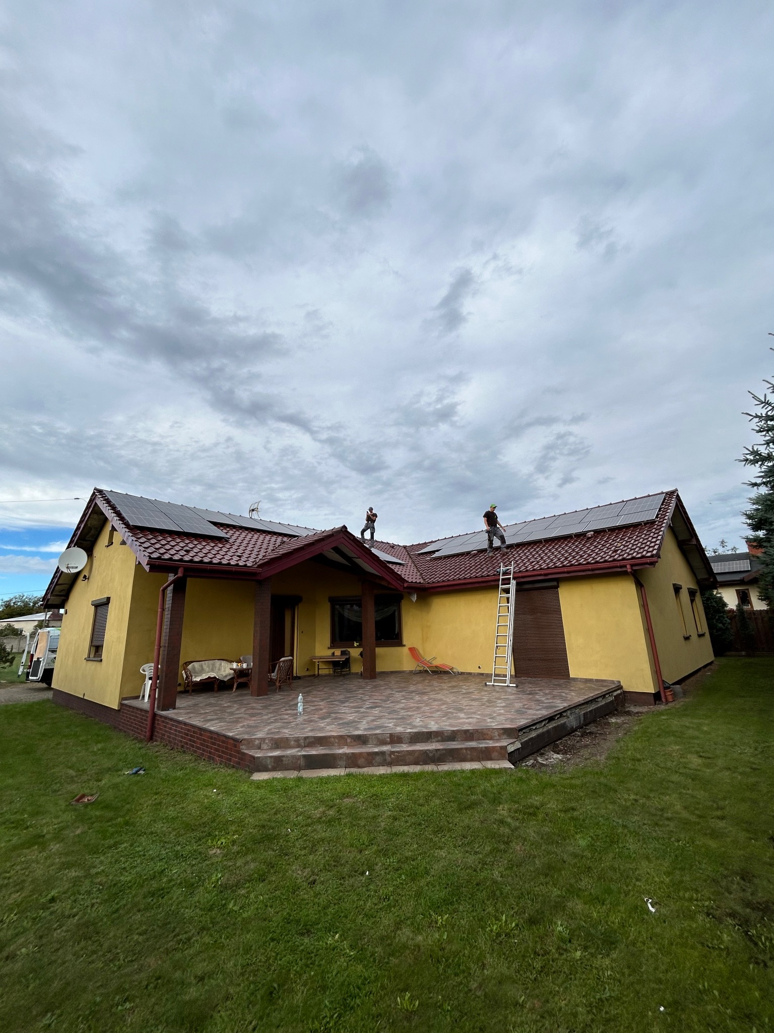 Żółty dom z panelami słonecznymi na dachu, montowanymi przez dwóch pracowników z użyciem drabiny. Pochmurne niebo w tle. Widok z poziomu trawnika.