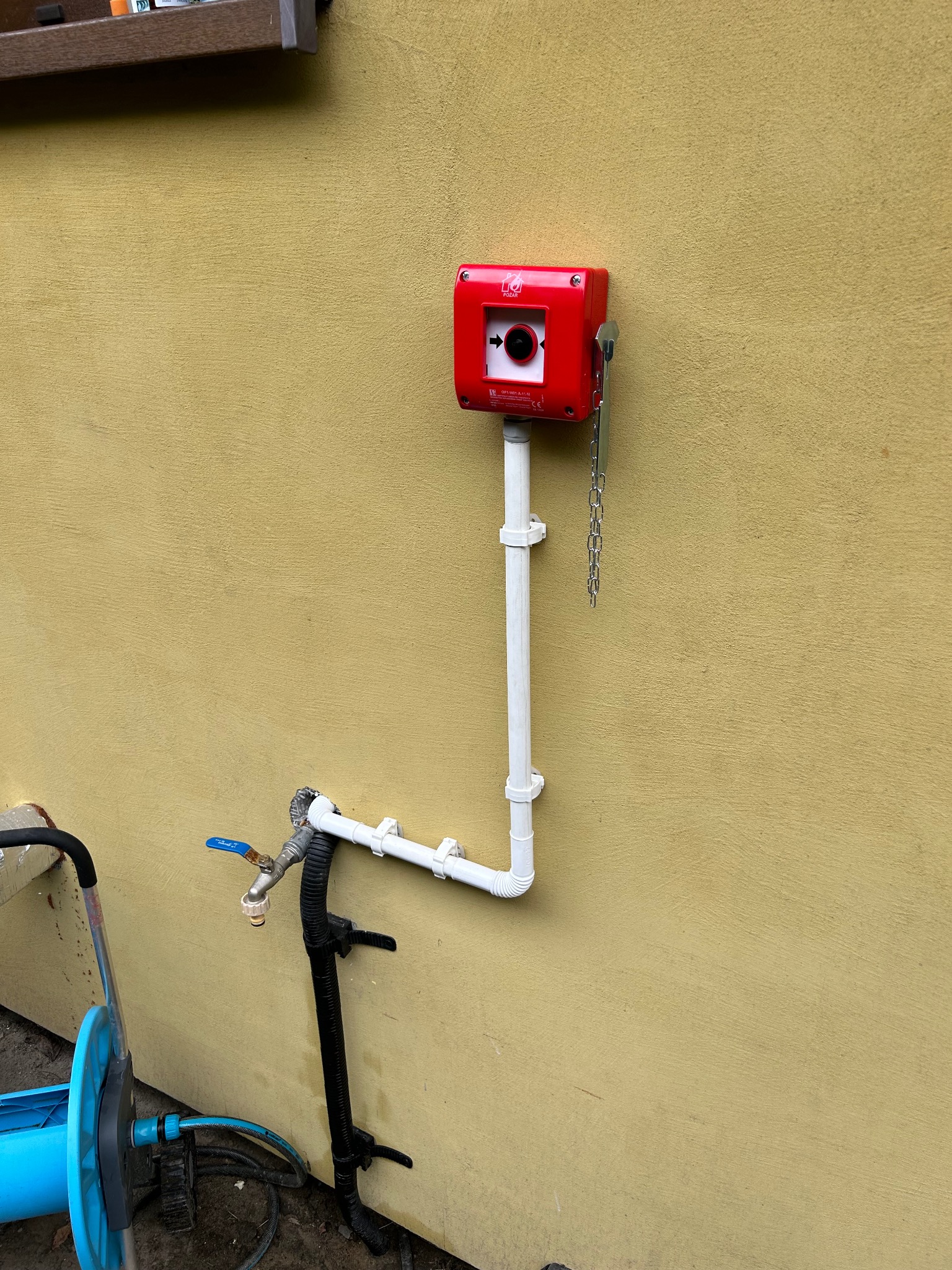 Czerwona skrzynka alarmowa z łańcuszkiem i rurami PCV na żółtej ścianie, obok kranu i zwiniętego węża ogrodowego. Ujęcie z bliska, dzienne światło.