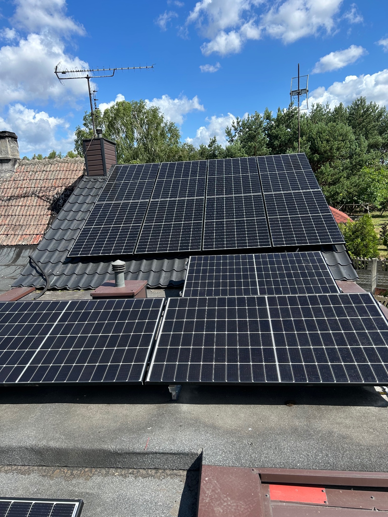 Panele fotowoltaiczne na dachu domu z anteną i kominem. Widok z dołu, błękitne niebo z chmurami w tle. Różne typy pokrycia dachowego.