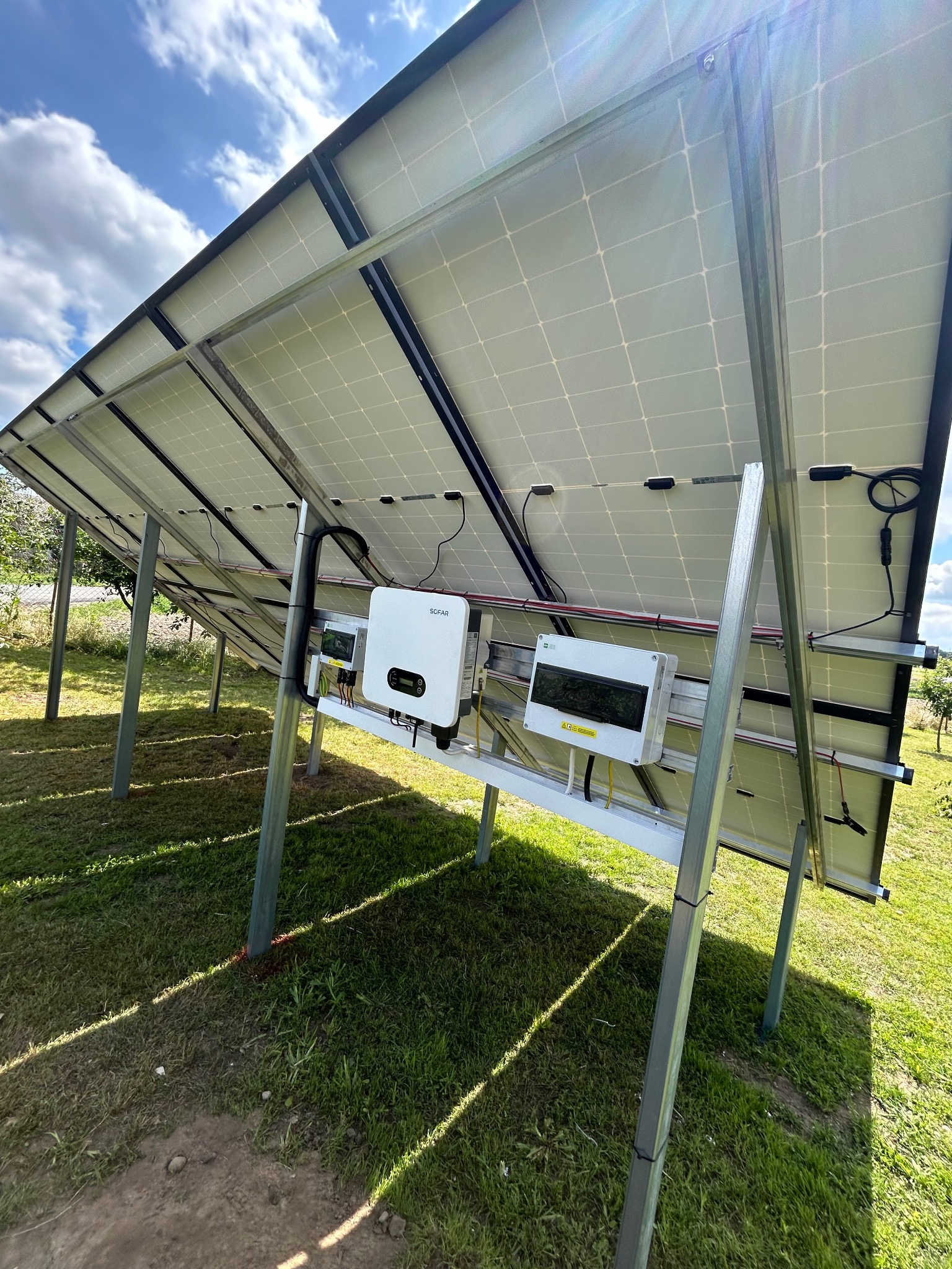 Montaż paneli słonecznych na konstrukcji wsporczej, widok od spodu z widocznym inwerterem Sofar i skrzynką zabezpieczeń na tle zielonej trawy i błękitnego nieba.