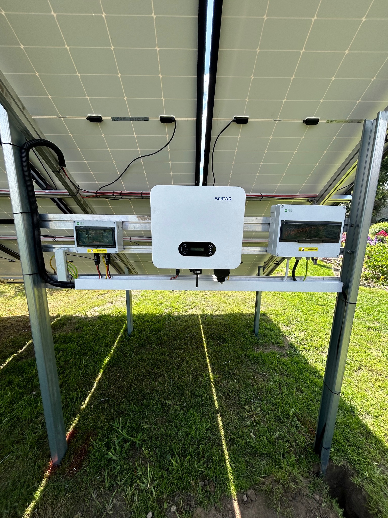 Montaż paneli fotowoltaicznych od spodu z widocznym inwerterem Sofar i skrzynkami elektrycznymi na metalowej konstrukcji, na tle trawnika i kwiatów.