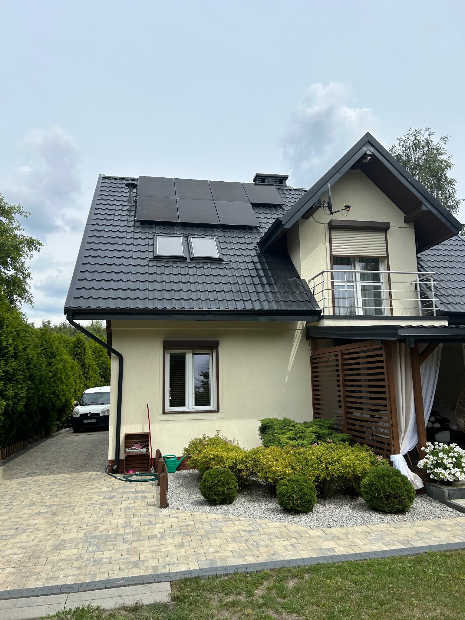 Dom jednorodzinny z panelami słonecznymi na dachu krytym grafitową dachówką. Widoczny balkon z metalową balustradą i zadbany ogród z kostką brukową.