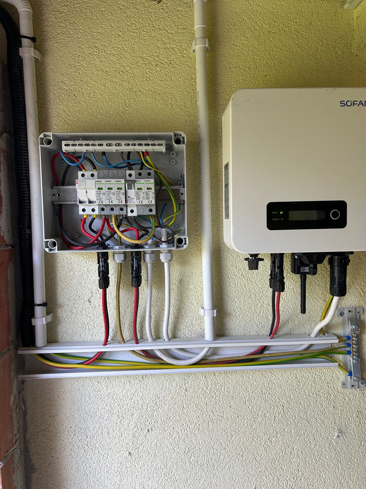 Instalacja fotowoltaiczna: zabezpieczenia w skrzynce, inwerter Sofar z podłączonymi kablami. Estetyczne prowadzenie przewodów w korytku. Ściana o fakturze baranka.