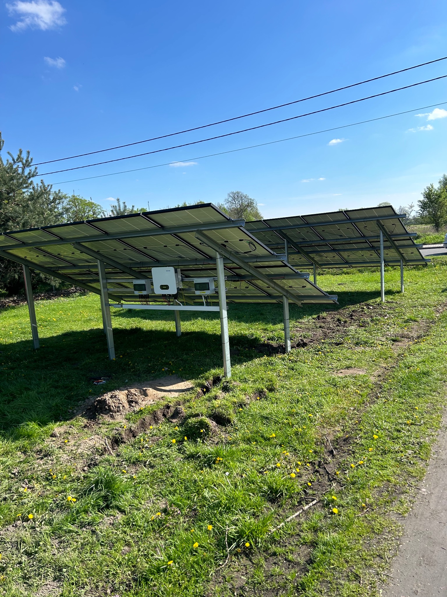 Instalacja paneli słonecznych na metalowej konstrukcji, widok z dołu. Widoczne falowniki i okablowanie. Trawa z żółtymi kwiatami w tle, błękitne niebo.