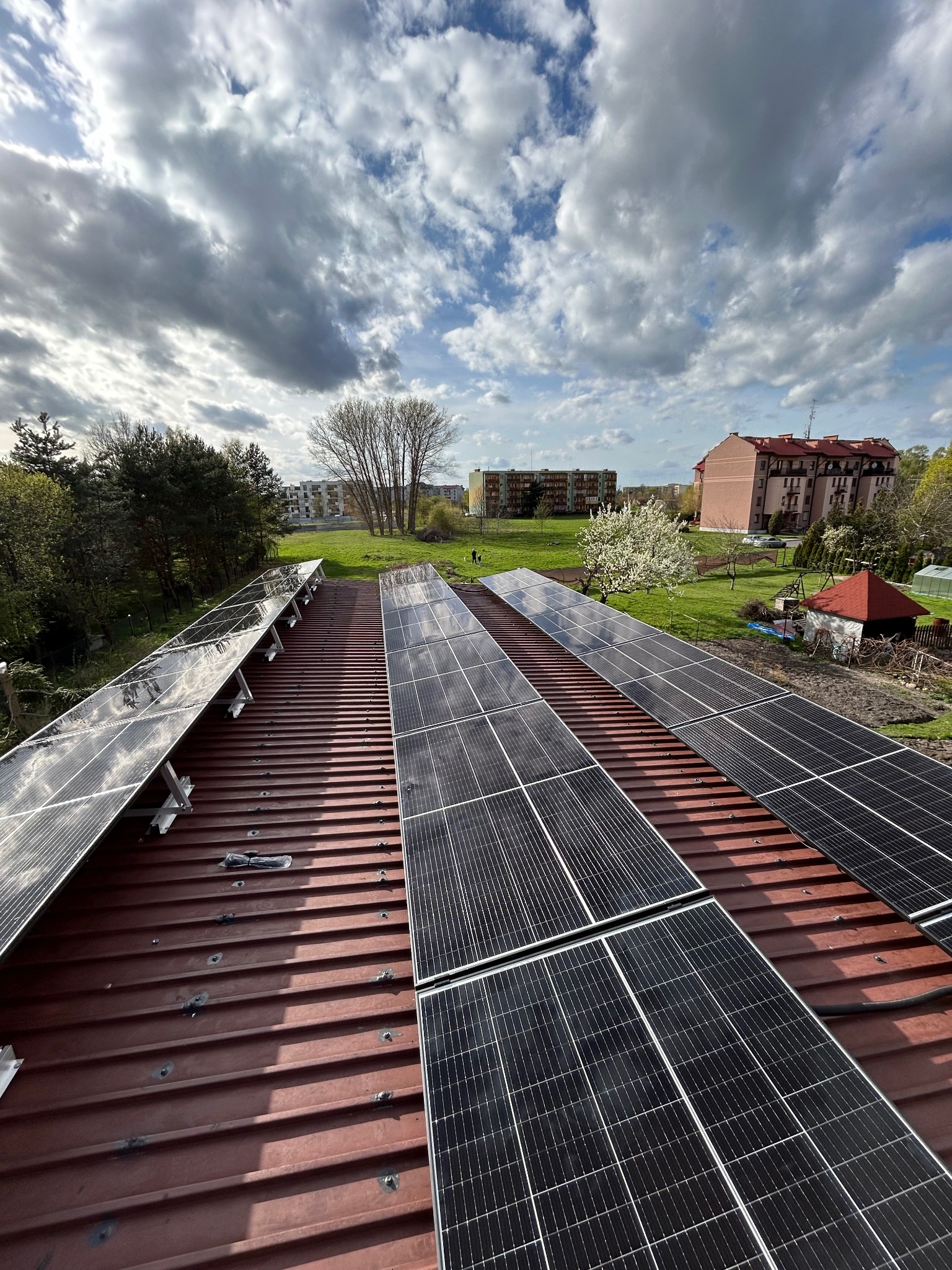 Panele słoneczne na dachu z blachy trapezowej, widok z góry na tle zabudowy miejskiej i pochmurnego nieba. Widoczne mocowania paneli.