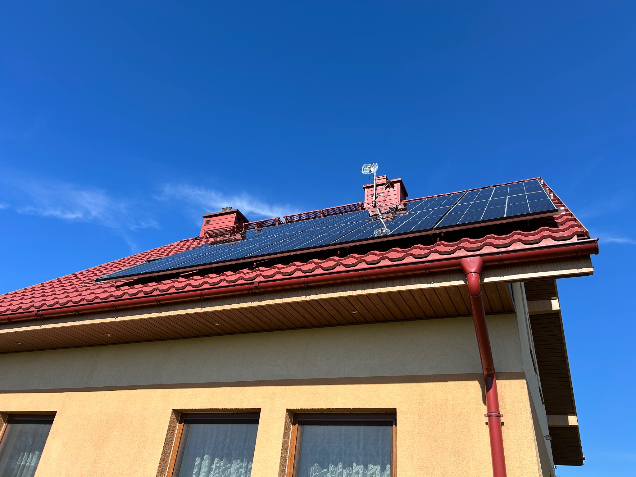 Panele słoneczne na dachu domu z czerwoną dachówką, widoczne okna i błękitne niebo. Instalacja w słoneczny dzień, antena na kominie.