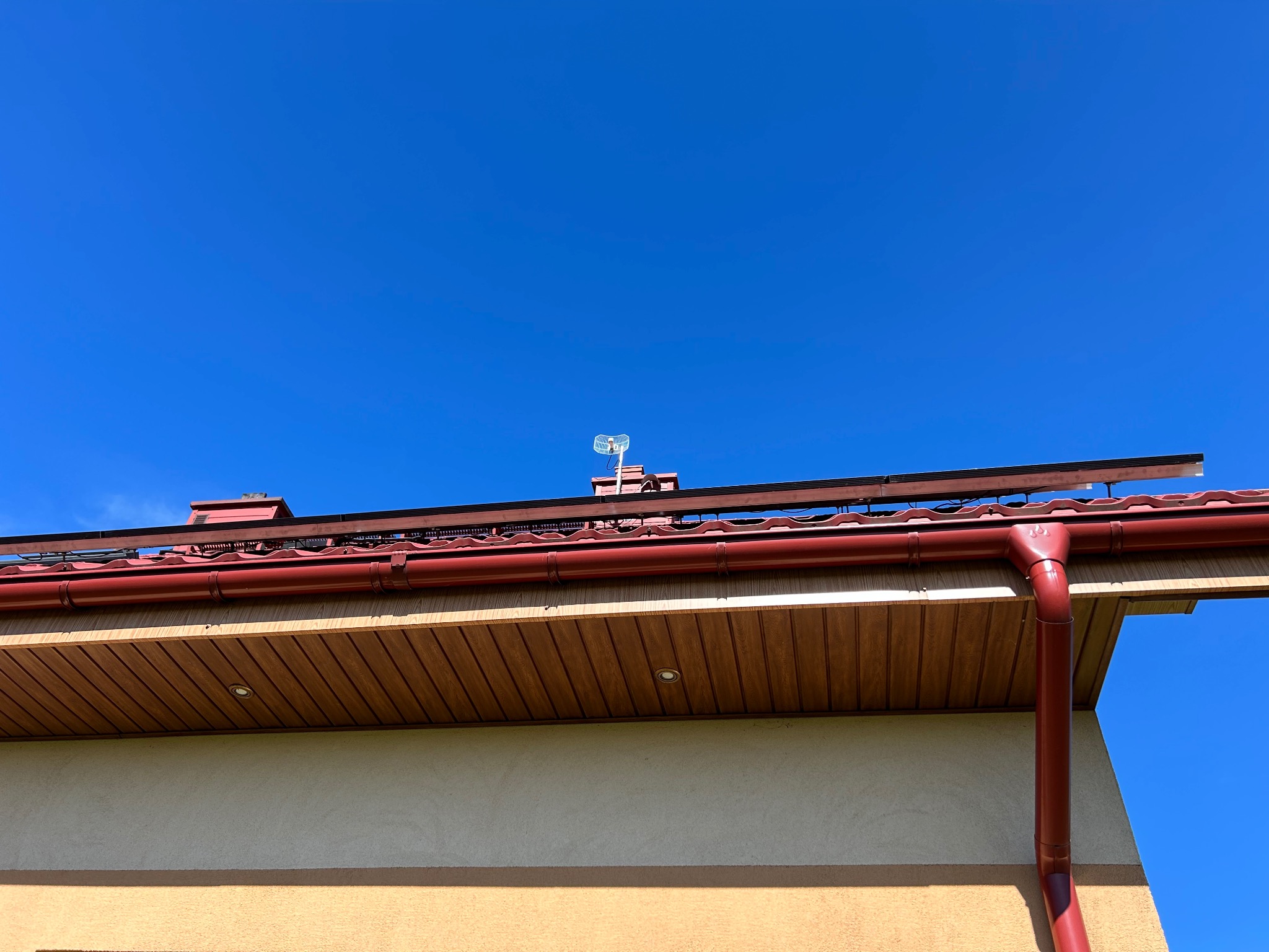 Czerwona metalowa rynna na elewacji domu z oświetleniem podbitki dachowej. Na dachu widoczna antena TV i instalacja solarna. Błękitne niebo w tle.