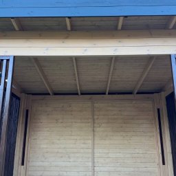 WOODCRAFTER ARTEM ZHYRNOV - Jasna, drewniana konstrukcja z ciemnymi, metalowymi elementami, widoczna od wewnątrz, z tłem niebieskiego nieba i zielonych drzew. Brukowana podłoga.