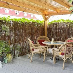 WOODCRAFTER ARTEM ZHYRNOV - Zadaszony taras z drewnianą pergolą, rattanowymi meblami i bambusową osłoną. Dekoracyjne oświetlenie żarówkowe. Szary taras z dywanem.