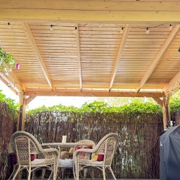 WOODCRAFTER ARTEM ZHYRNOV - Zadaszony taras z drewnianą konstrukcją, oświetleniem girlandą żarówkową i meblami z rattanu. W tle widoczna bambusowa osłona i zieleń.