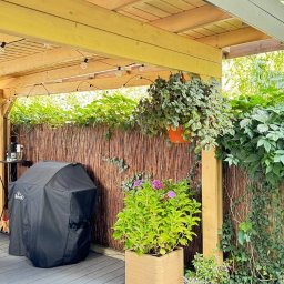 WOODCRAFTER ARTEM ZHYRNOV - Zadaszony taras z drewnianą pergolą, oświetleniem i roślinami w donicach. Grill pod pokrowcem, mata na podłodze, kamyki dekoracyjne. Urokliwe miejsce do relaksu.
