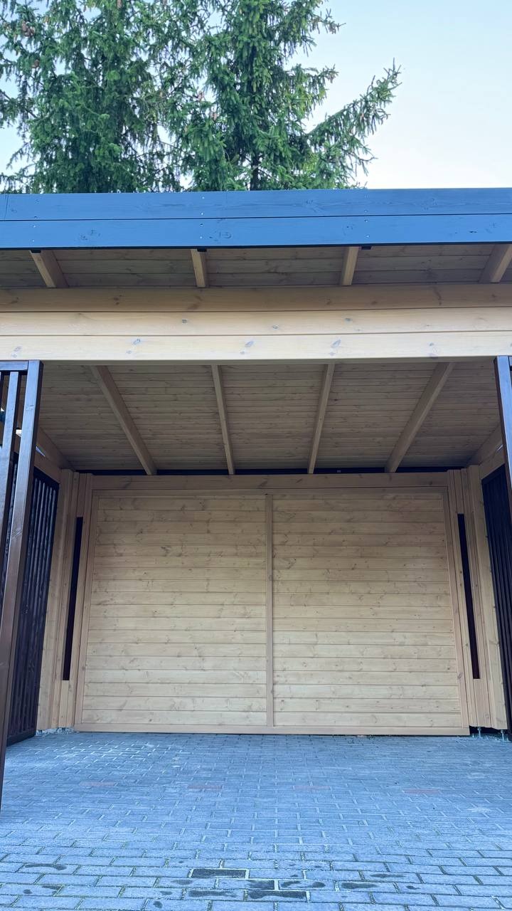 Jasna, drewniana konstrukcja z ciemnymi, metalowymi elementami, widoczna od wewnątrz, z tłem niebieskiego nieba i zielonych drzew. Brukowana podłoga.