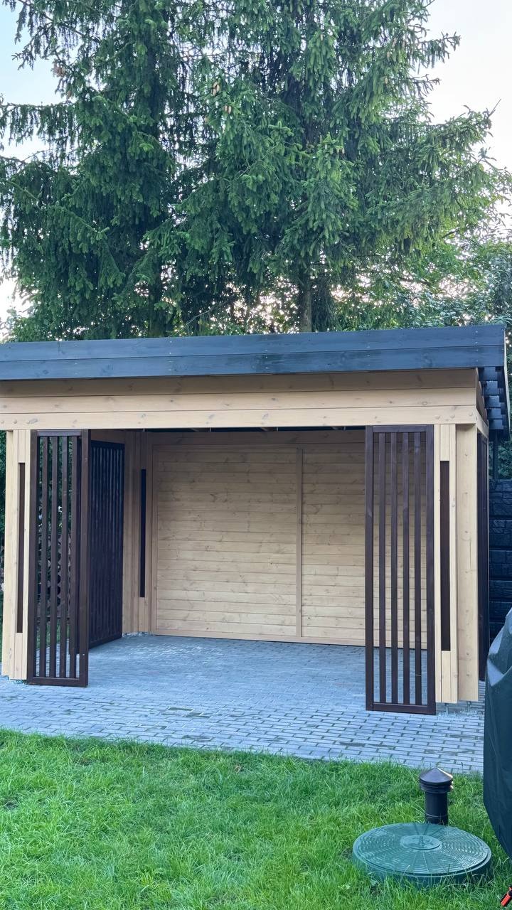 Drewniana altana z płaskim dachem i ażurowymi panelami wejściowymi, na tle zielonego trawnika i drzew. Widok z przodu, stonowana kolorystyka.