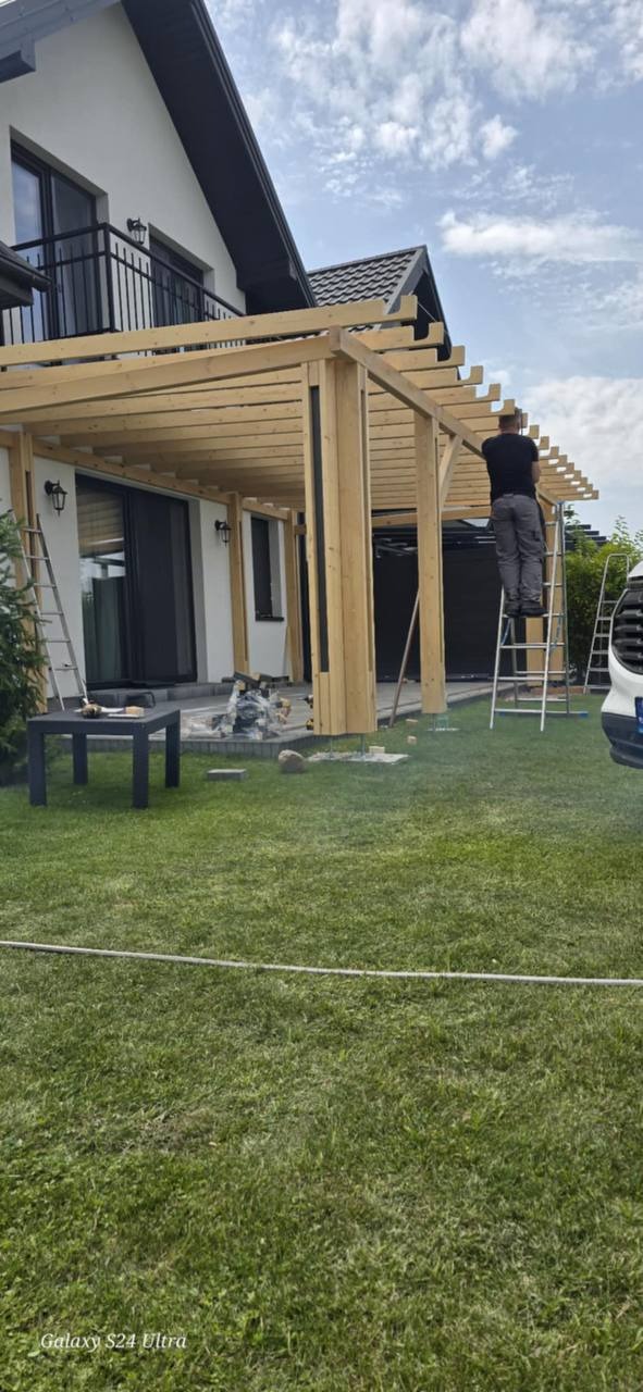 Montaż drewnianej pergoli przy domu z balkonem. Pracownik na drabinie mocuje elementy konstrukcji. Widoczne narzędzia i materiały budowlane na trawniku.