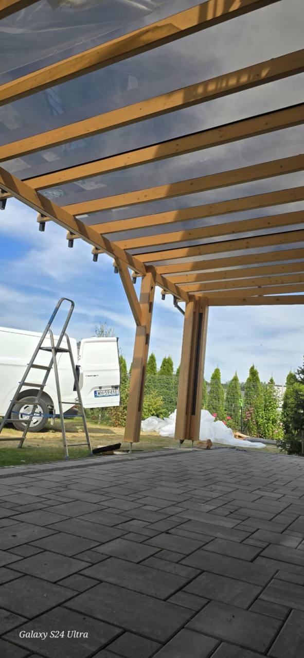 Drewniana pergola z przezroczystym zadaszeniem, widok od dołu, drabina oparta o konstrukcję, częściowo widoczny biały bus. Elementy montażowe.